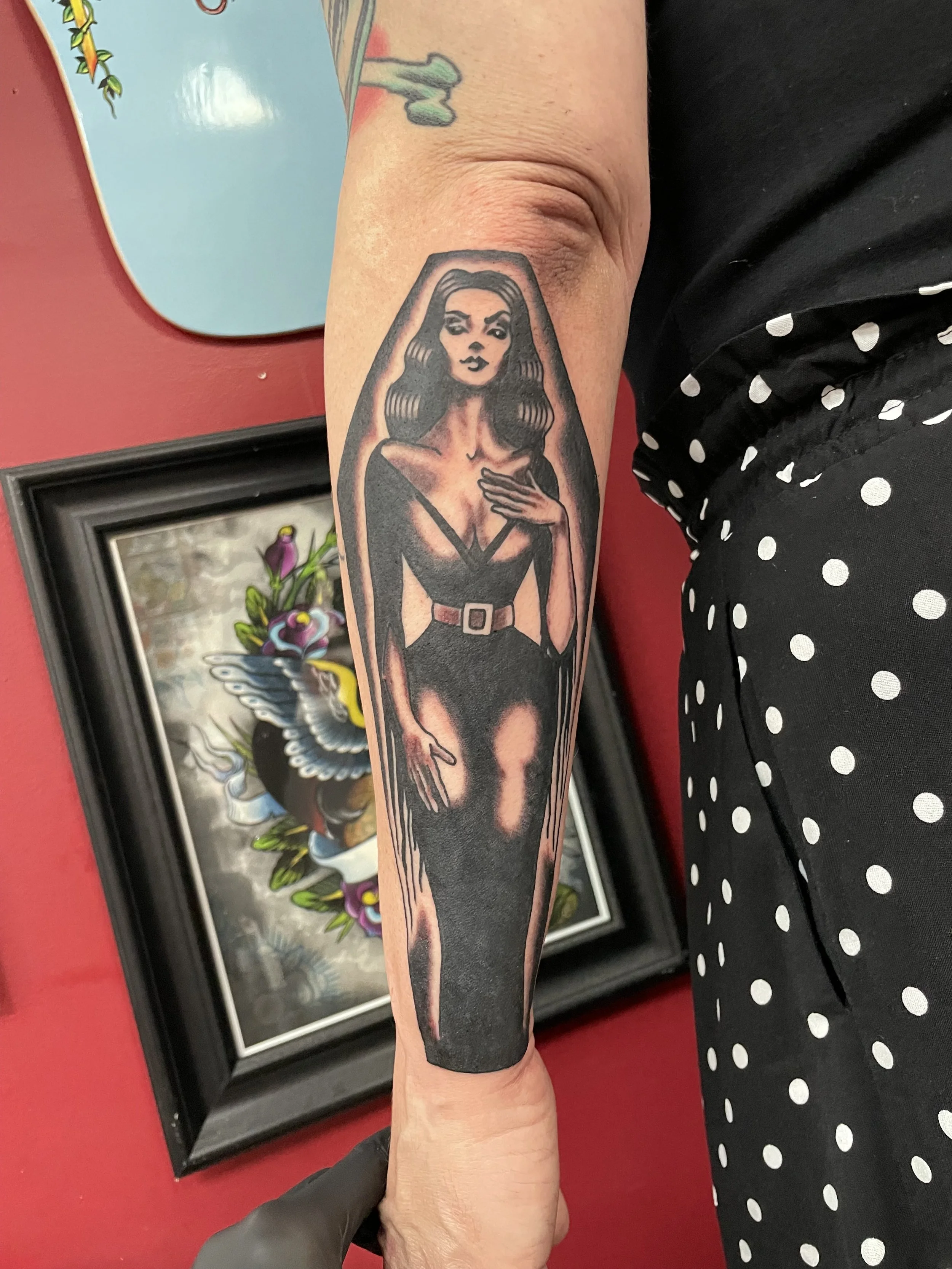 Black and grey forearm tattoo featuring a classic horror-inspired pinup woman in a form-fitting black gown with a cinched belt and dramatic long dark hair, framed within a bold coffin silhouette, custom tattoo by Nick at Big Kahuna Tattoo in Boca Rat