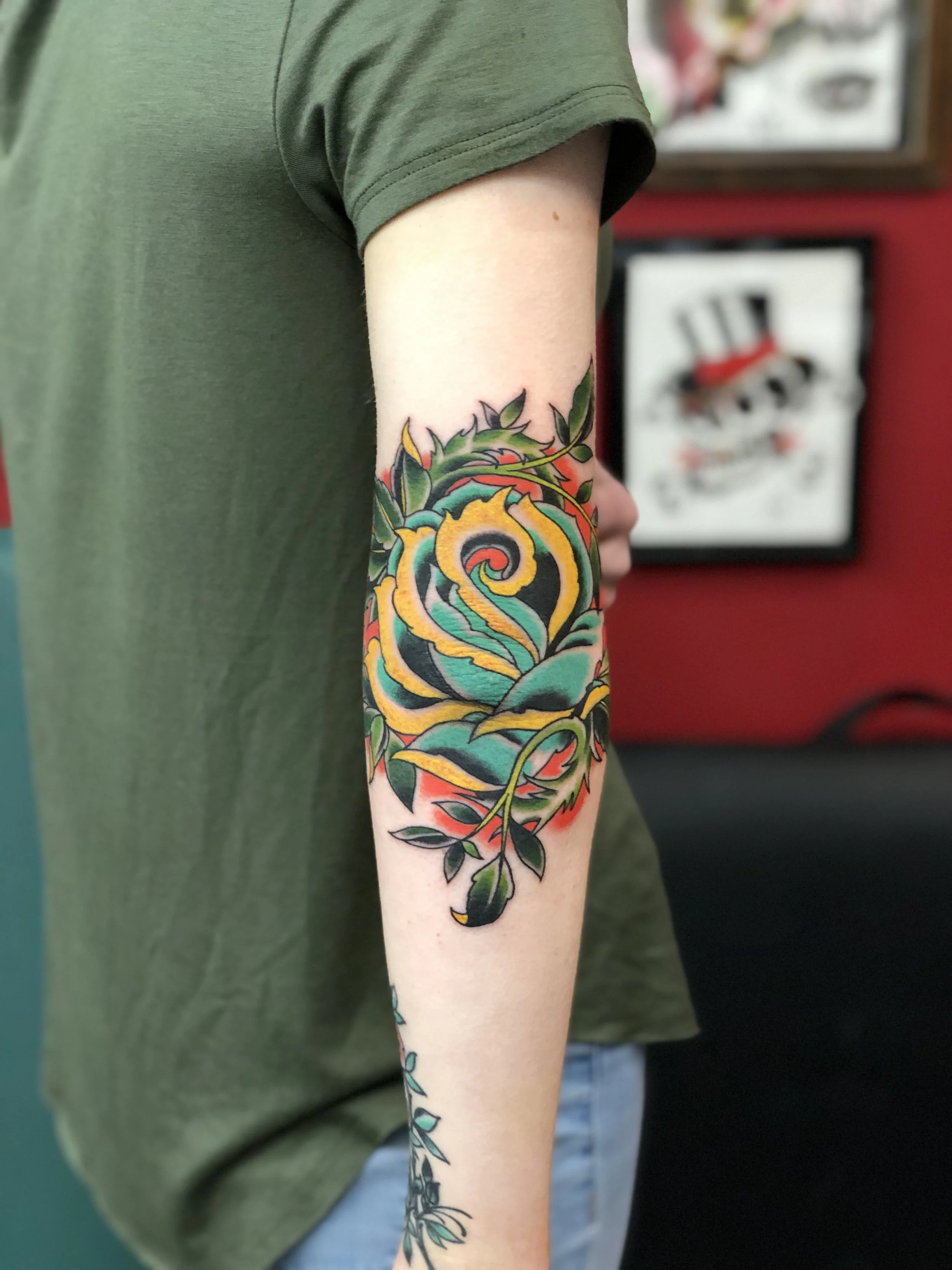 Bold traditional American color elbow tattoo featuring a large teal and yellow rose with green thorny vines and red accents, custom tattoo by Nick at Big Kahuna Tattoo in Boca Raton, FL.