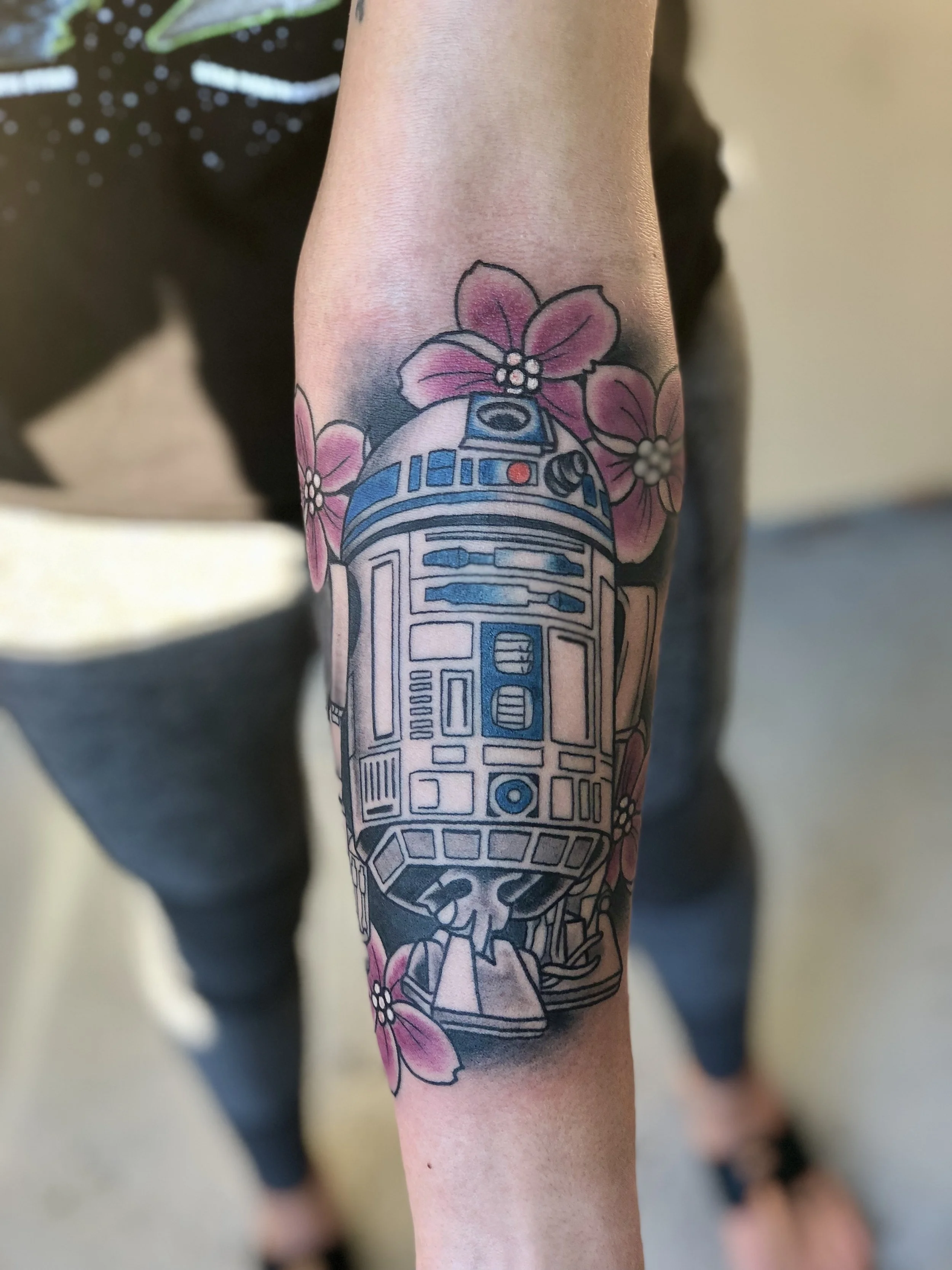 Color forearm tattoo featuring R2-D2 from Star Wars surrounded by soft pink cherry blossoms, blending sci-fi and floral elements, custom tattoo by Nick at Big Kahuna Tattoo in Boca Raton, FL.