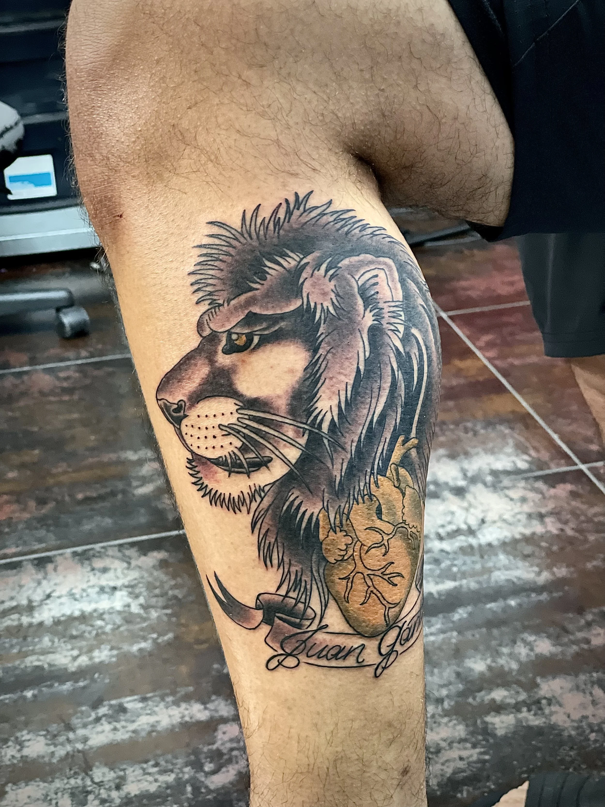 A lion in side profile with a wild flowing mane, and a golden anatomical heart, with the name "Juan" scrolled across a banner below. The kind of personal piece that means something every time you look at it. Black and grey shin tattoo by Nick at Big 