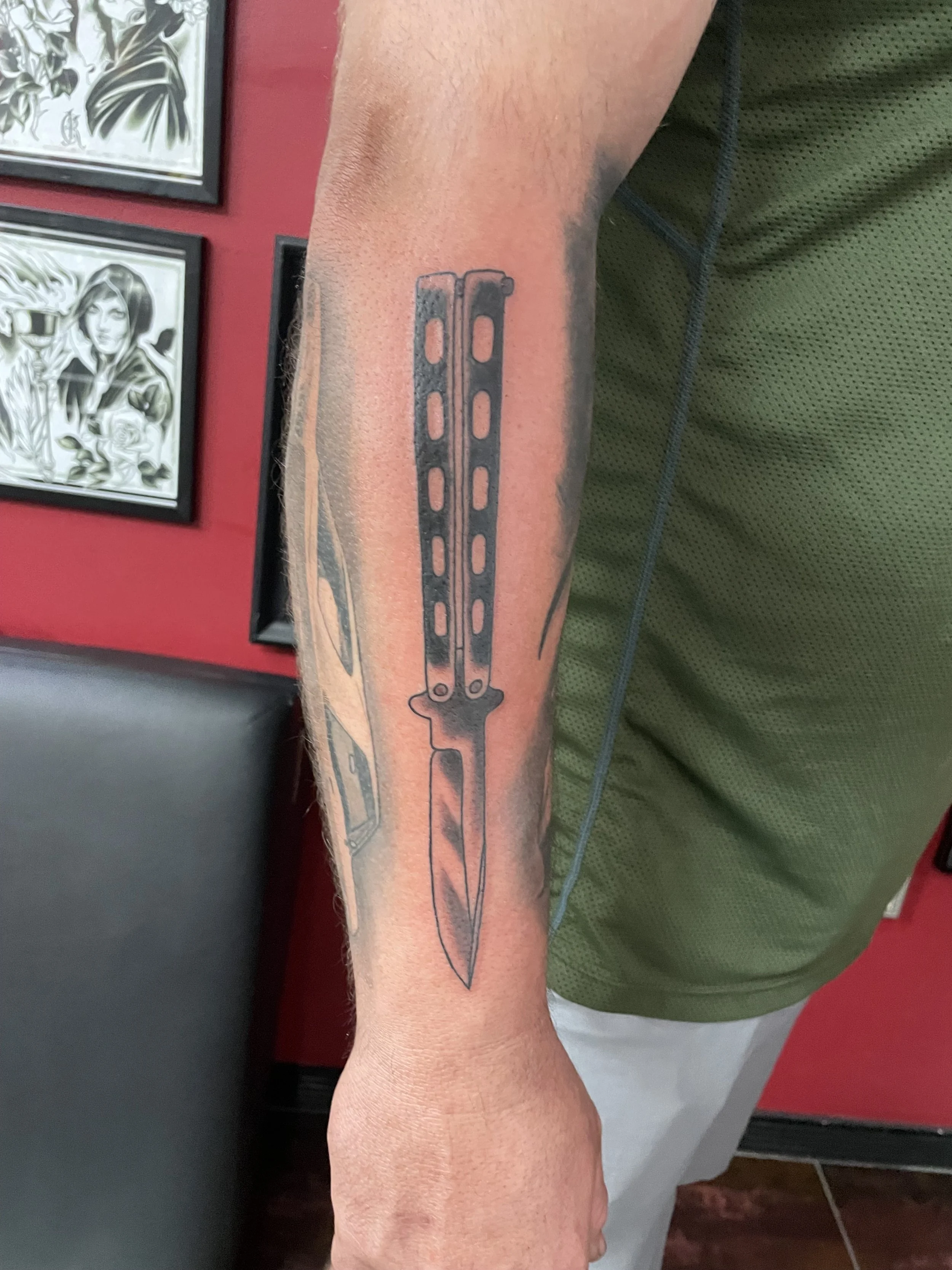 Black and grey forearm tattoo featuring a detailed balisong butterfly knife with perforated handles and a long tapered blade, rendered with precise shading and metallic detail, custom tattoo by Nick at Big Kahuna Tattoo in Boca Raton, FL.