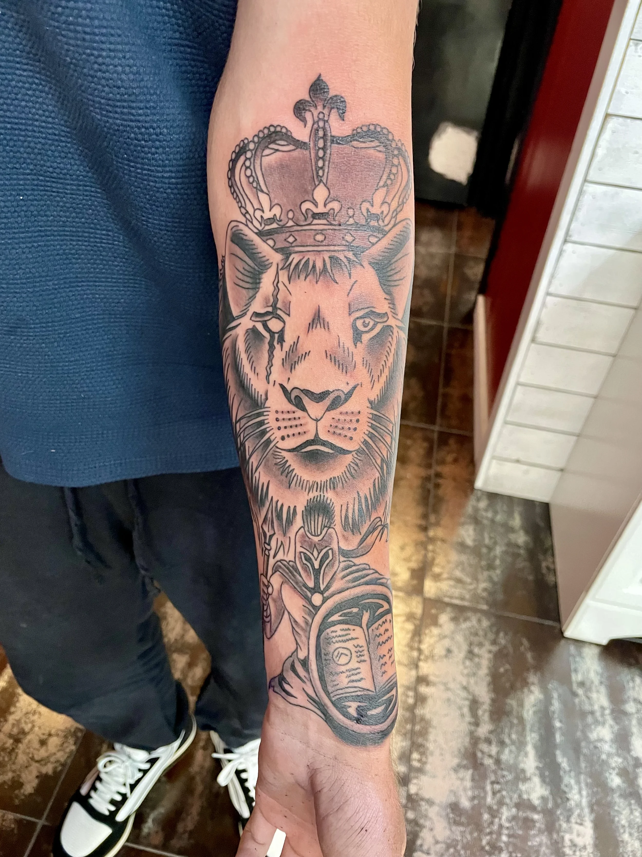 Black and grey forearm tattoo featuring a regal front-facing lion wearing an ornate royal crown, with a small armored Spartan warrior figure holding a shield that depicts an open book below, custom tattoo by Nick at Big Kahuna Tattoo in Boca Raton, F