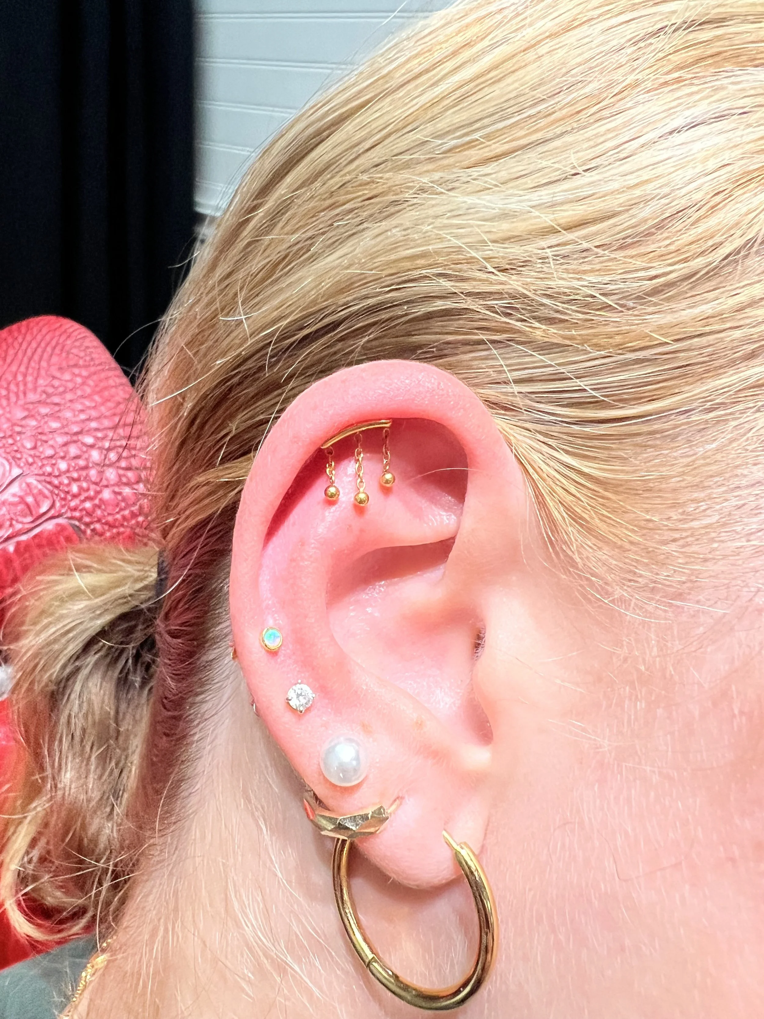 
Curated ear with a gold rook piercing featuring a dangle chain with gold bead drops, opal tragus stud, crystal lobe stud, pearl lobe stud, and a chunky gold hoop. Delicate and feminine gold stack. Done at Big Kahuna Tattoo in Boca Raton.