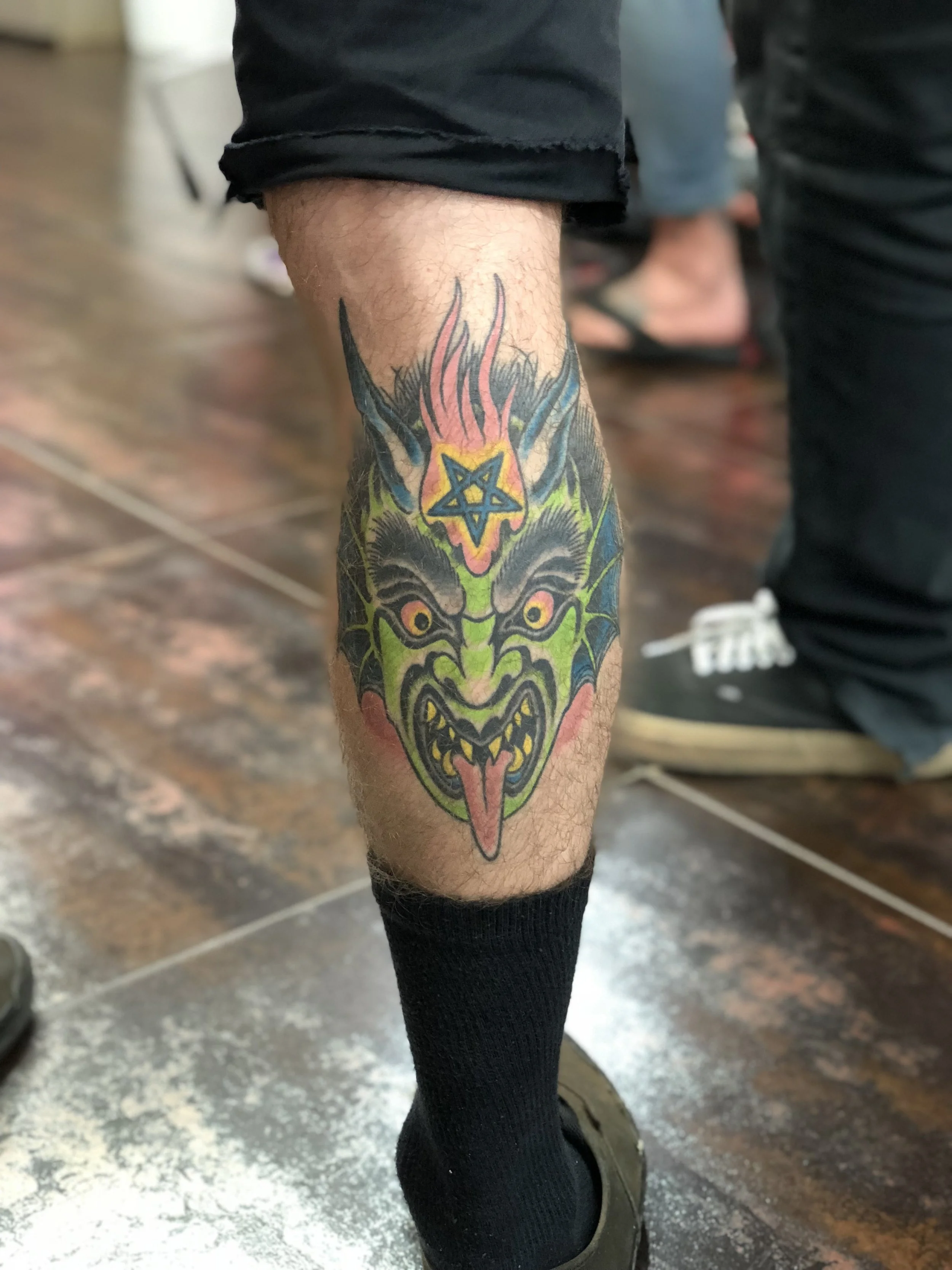Traditional American color tattoo featuring a green devil face with horns, bat wings, a pentagram, and pink flames on the calf, bold and vibrant custom work by Nick at Big Kahuna Tattoo in Boca Raton, FL.