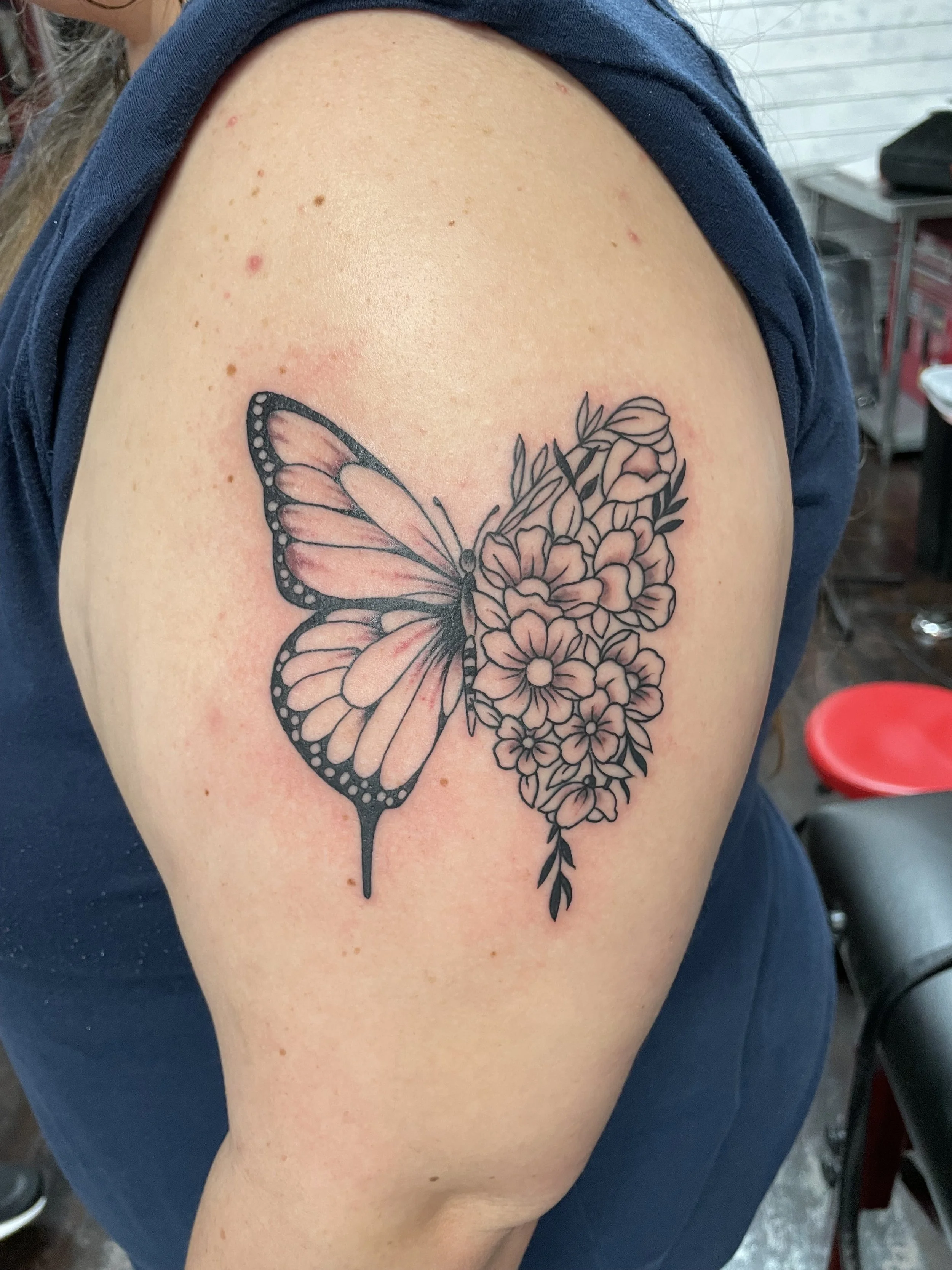 Fine Line black and grey shoulder tattoo featuring a split design with one side showing a monarch butterfly with soft pink shading and the other side transforming into a bouquet of detailed flowers and leaves, custom tattoo by Nick at Big Kahuna Tatt