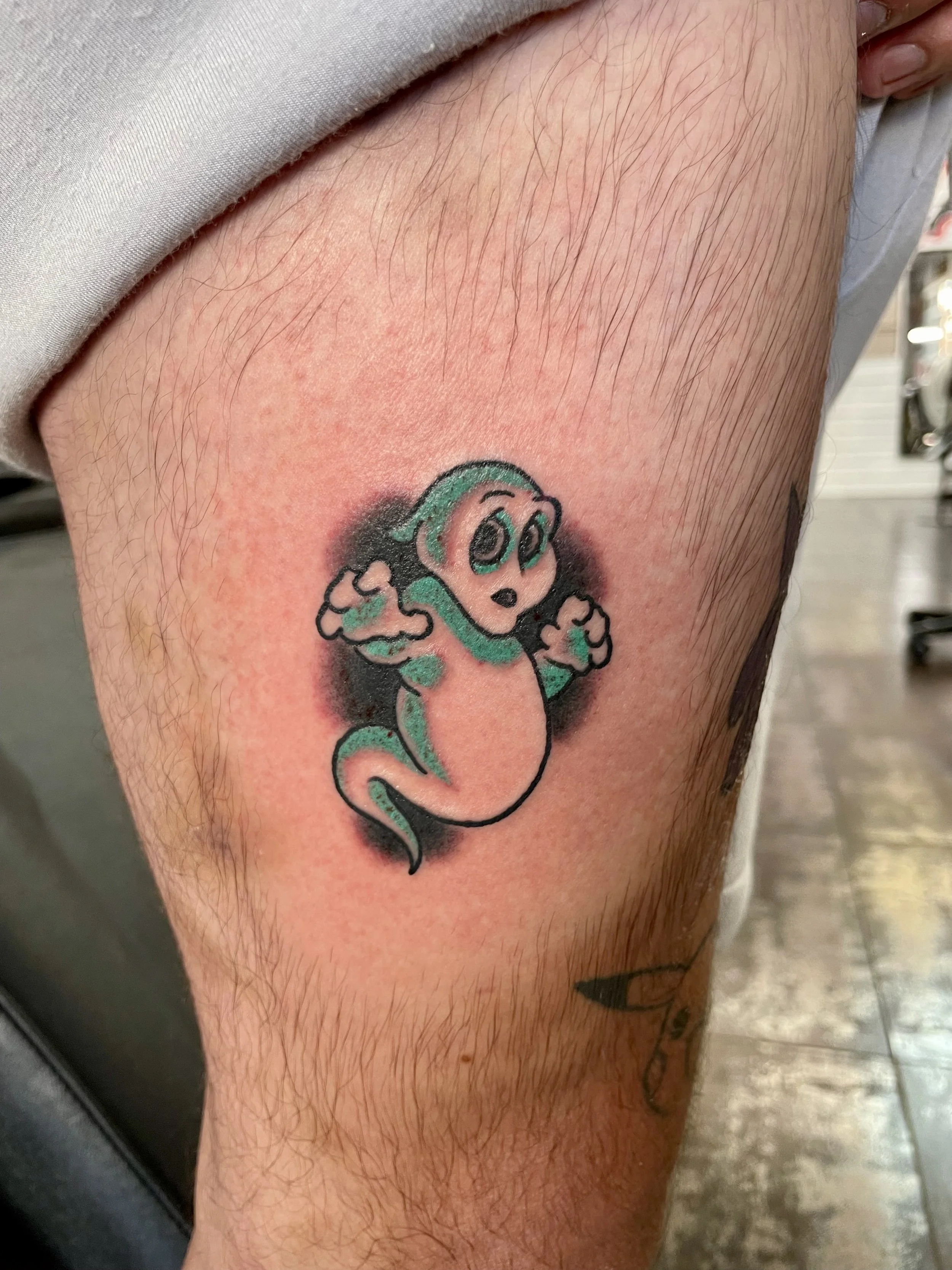 Color leg tattoo featuring a friendly cartoon ghost in teal and white with wide expressive eyes and raised hands, floating against a soft dark shadow background, custom tattoo by Nick at Big Kahuna Tattoo in Boca Raton, FL.