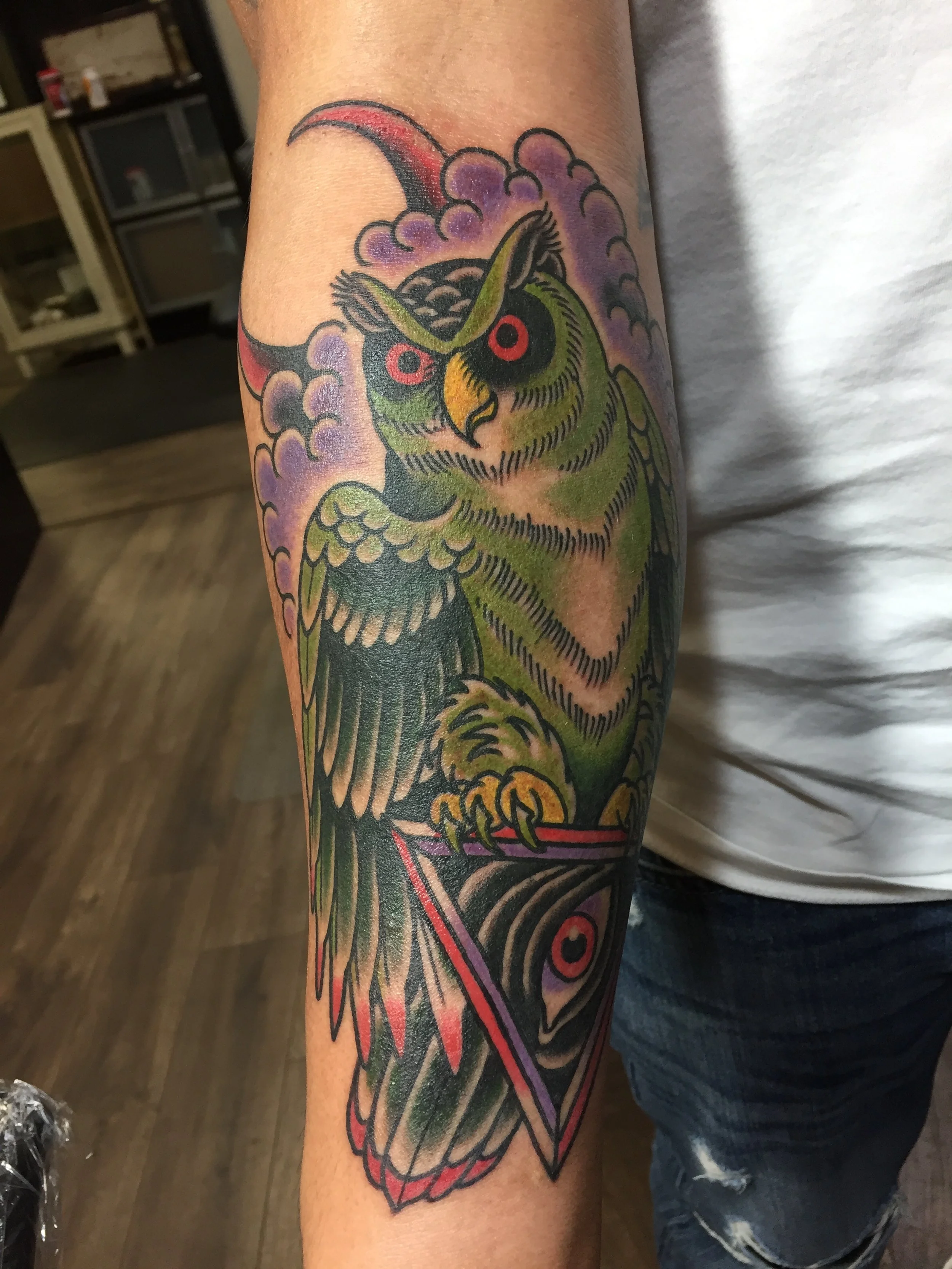 Traditional American color forearm tattoo featuring a green horned owl with red eyes clutching an all-seeing eye triangle in its talons, surrounded by purple clouds and a red crescent moon that echoes the eye motif throughout the design, custom tatto