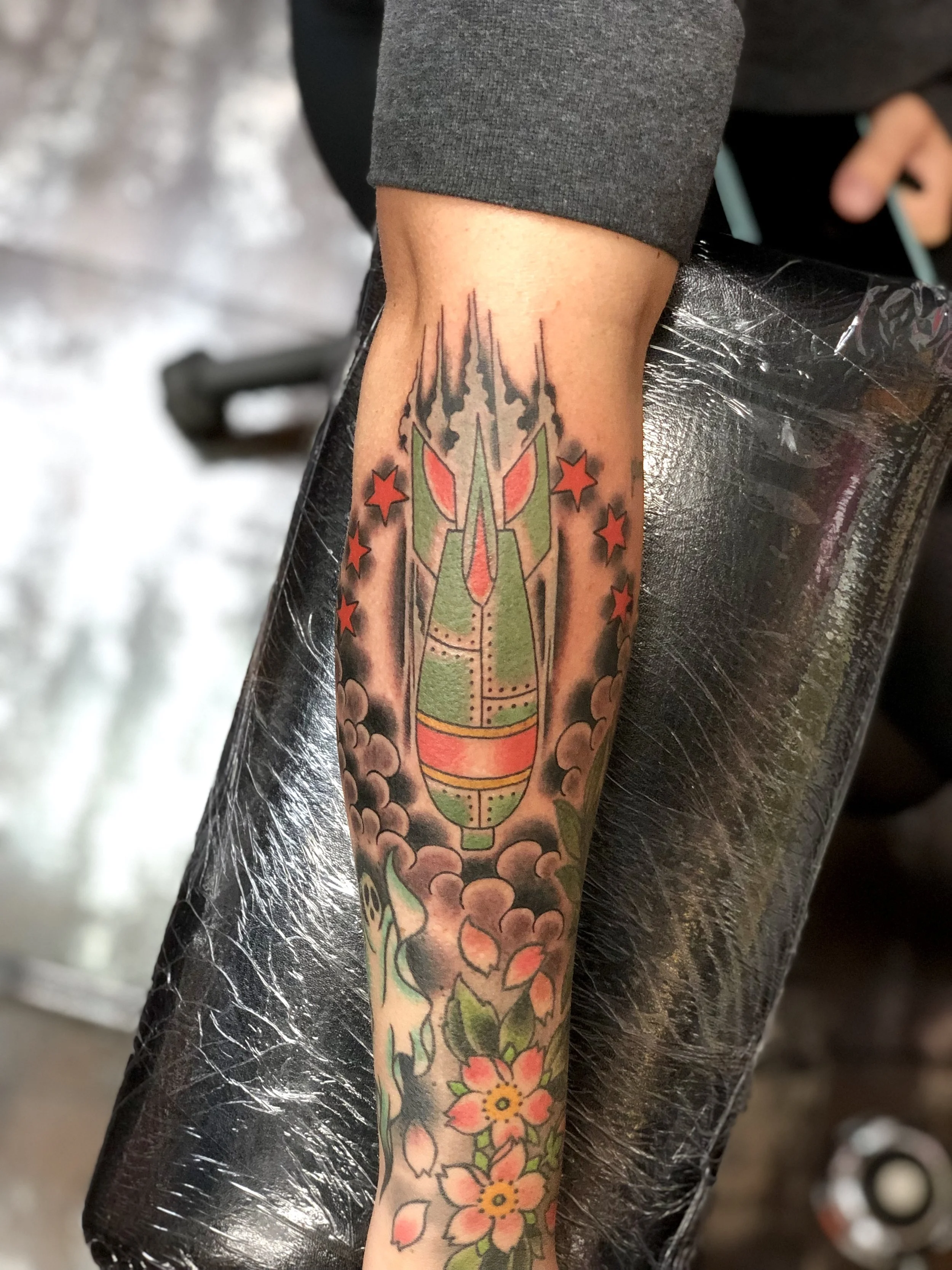 Traditional American color forearm tattoo featuring a green military bomb launching upward through smoke and red stars, surrounded by cherry blossoms, custom tattoo by Nick at Big Kahuna Tattoo in Boca Raton, FL.