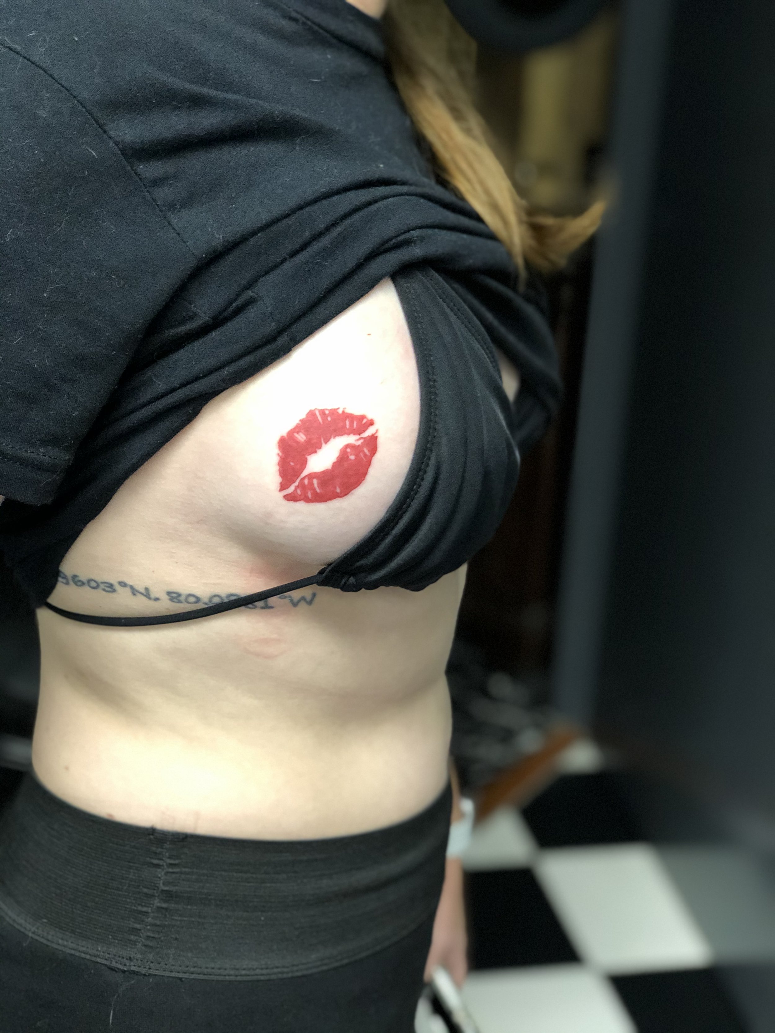 Minimalist rib tattoo combining a bold red lipstick kiss mark with fine line GPS coordinate text, custom tattoo by Nick at Big Kahuna Tattoo in Boca Raton, FL.