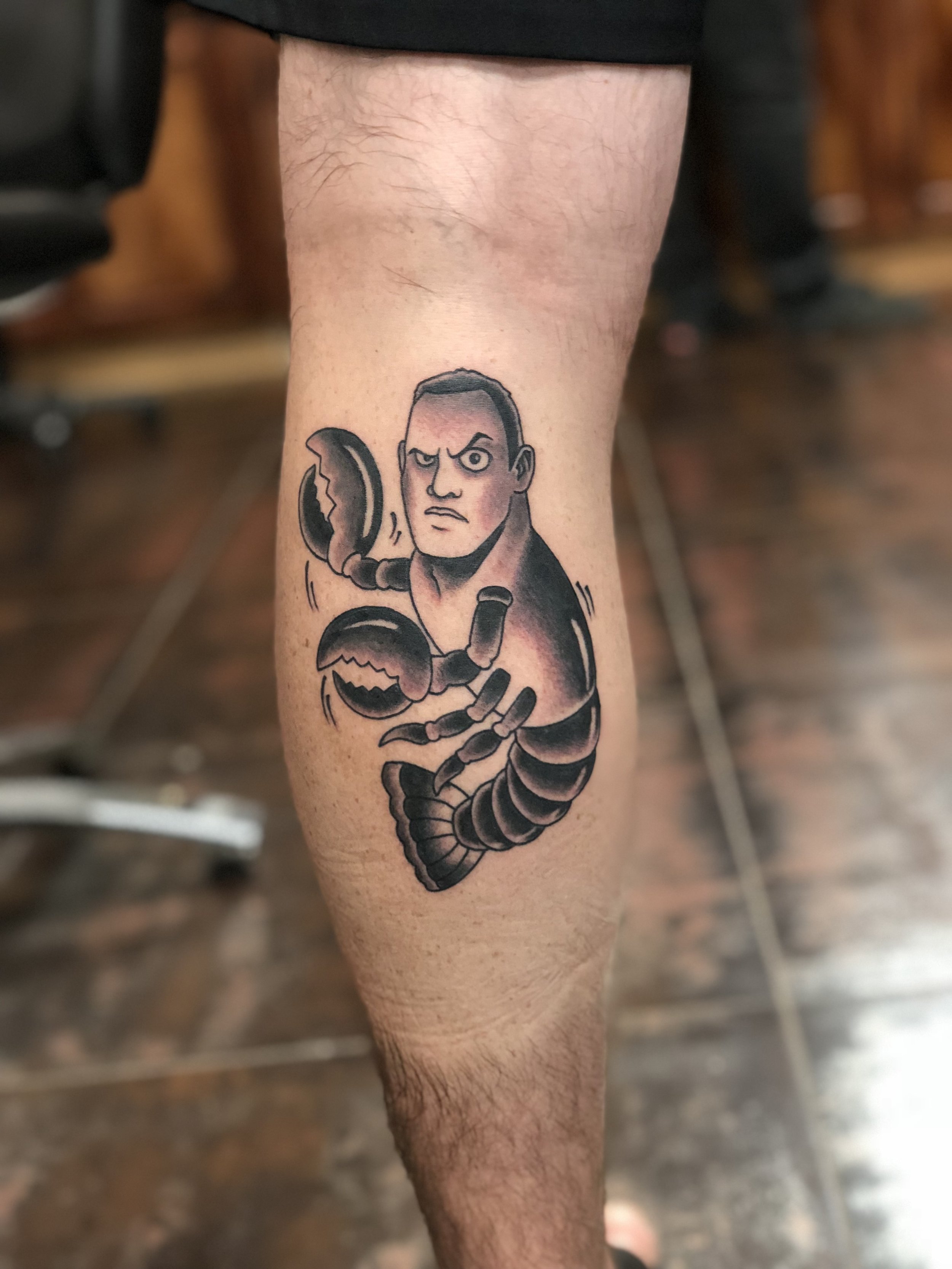 Humorous traditional American black and grey calf tattoo featuring a cartoon caricature of Dwayne "The Rock" Johnson as a lobster, a playful visual pun on the classic B-52's song "Rock Lobster," custom tattoo by Nick at Big Kahuna Tattoo in Boca Rato