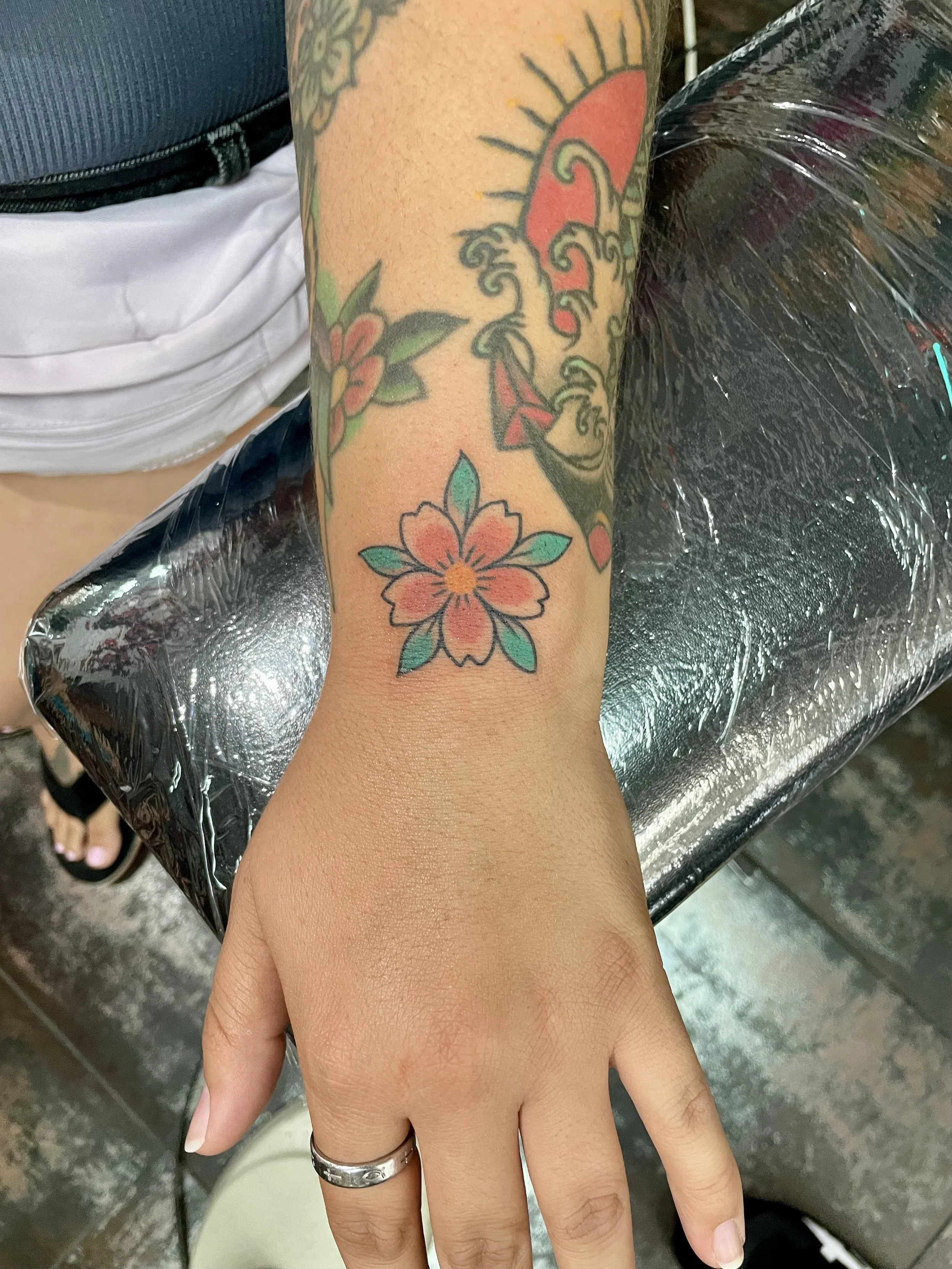 Color Small color wrist tattoo featuring a single cherry blossom flower with soft pink petals, a gold center, and teal green leaves, custom tattoo by Nick at Big Kahuna Tattoo in Boca Raton, FL.