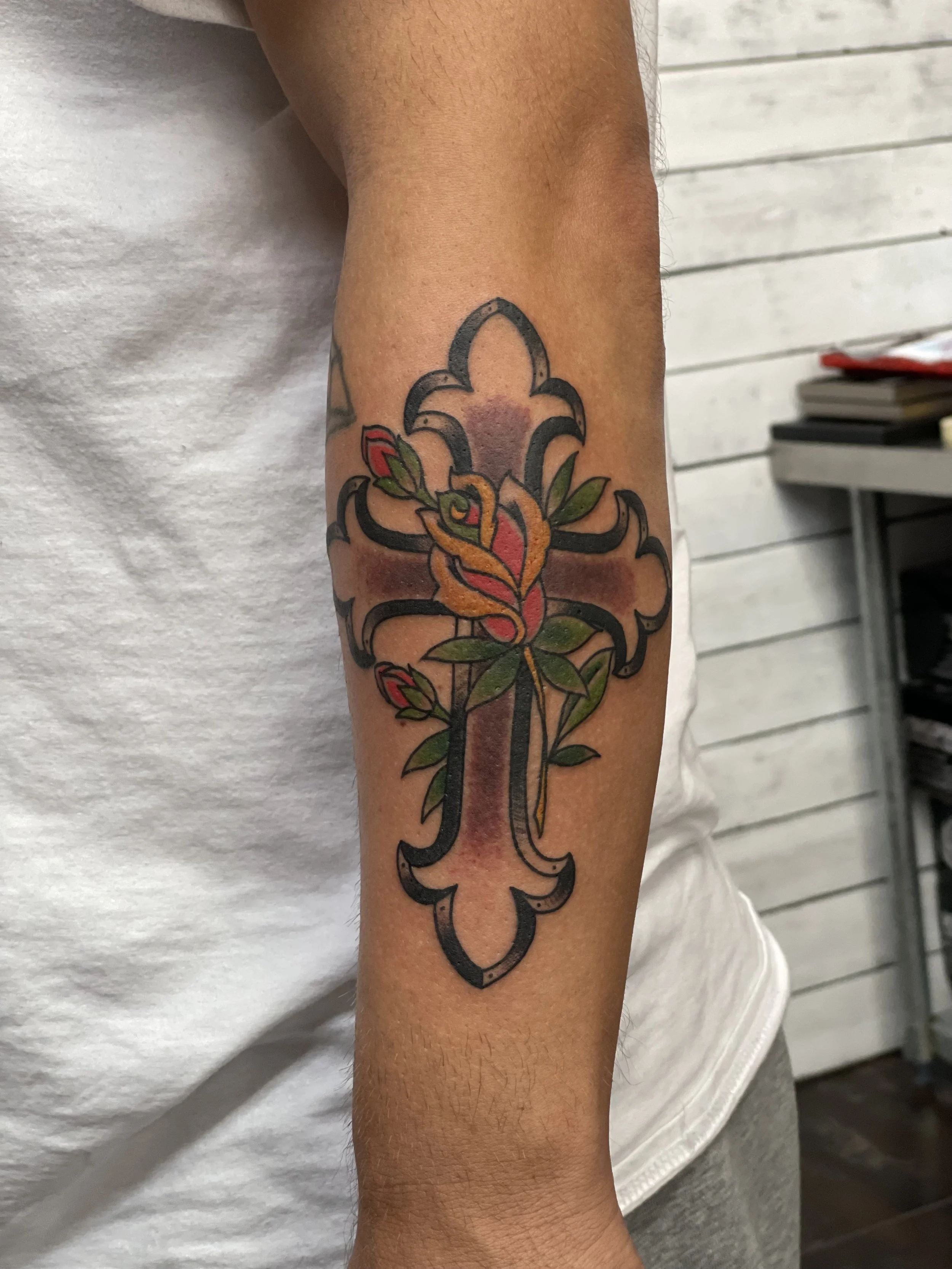 Traditional American color forearm tattoo featuring an ornate cross with flared ends and a blooming rose at the center, accented with red buds and green leaves, custom tattoo by Nick at Big Kahuna Tattoo in Boca Raton, FL.