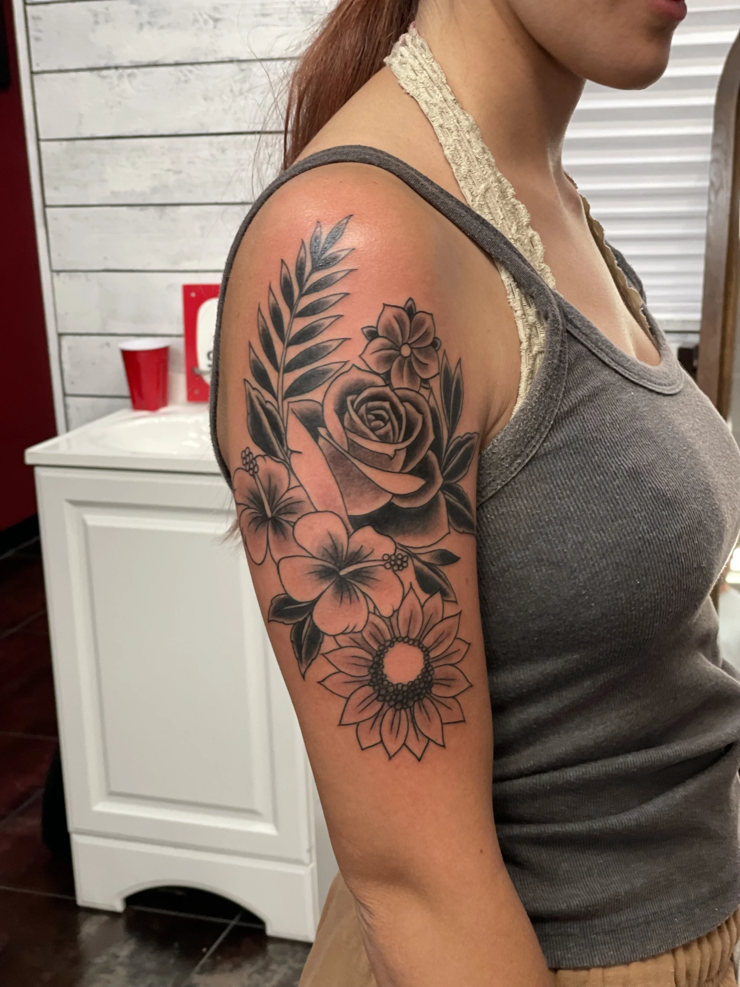 Fine line black and grey upper arm tattoo featuring a lush floral bouquet with a large rose at center, surrounded by a sunflower, hibiscus blossoms, small wildflowers, and botanical fronds, custom tattoo by Nick at Big Kahuna Tattoo in Boca Raton, FL