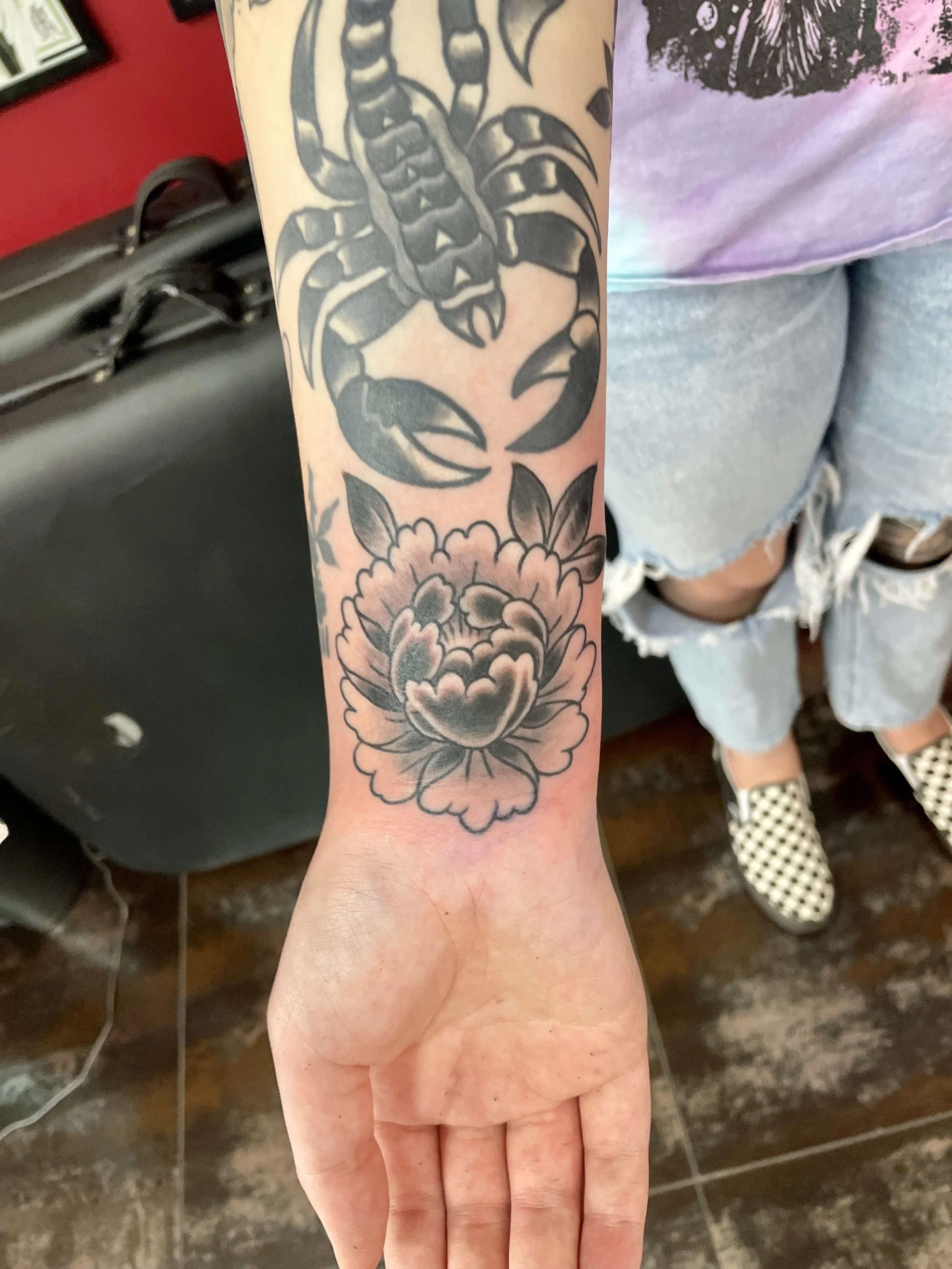 Black and grey wrist tattoo featuring a large fully bloomed peony with layered petals and dark leaf accents, positioned below existing scorpion tattoo, custom tattoo by Nick at Big Kahuna Tattoo in Boca Raton, FL.