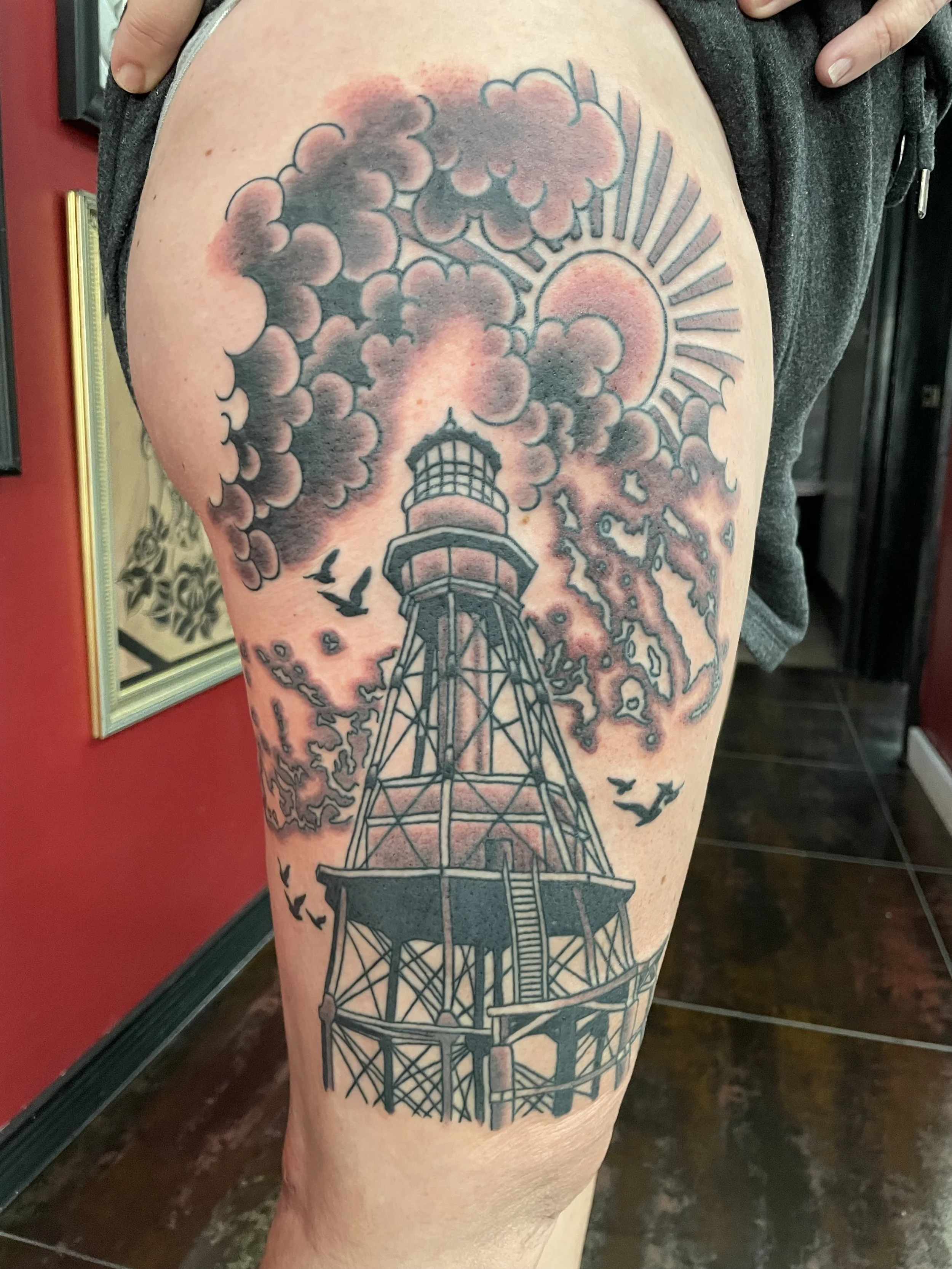 Black and grey thigh tattoo featuring a tall detailed iron-frame lighthouse inspired by the Florida Keys, rising into a dramatic sky of billowing clouds and a radiant sun with bold rays, with birds in flight on either side, custom tattoo by Nick at B