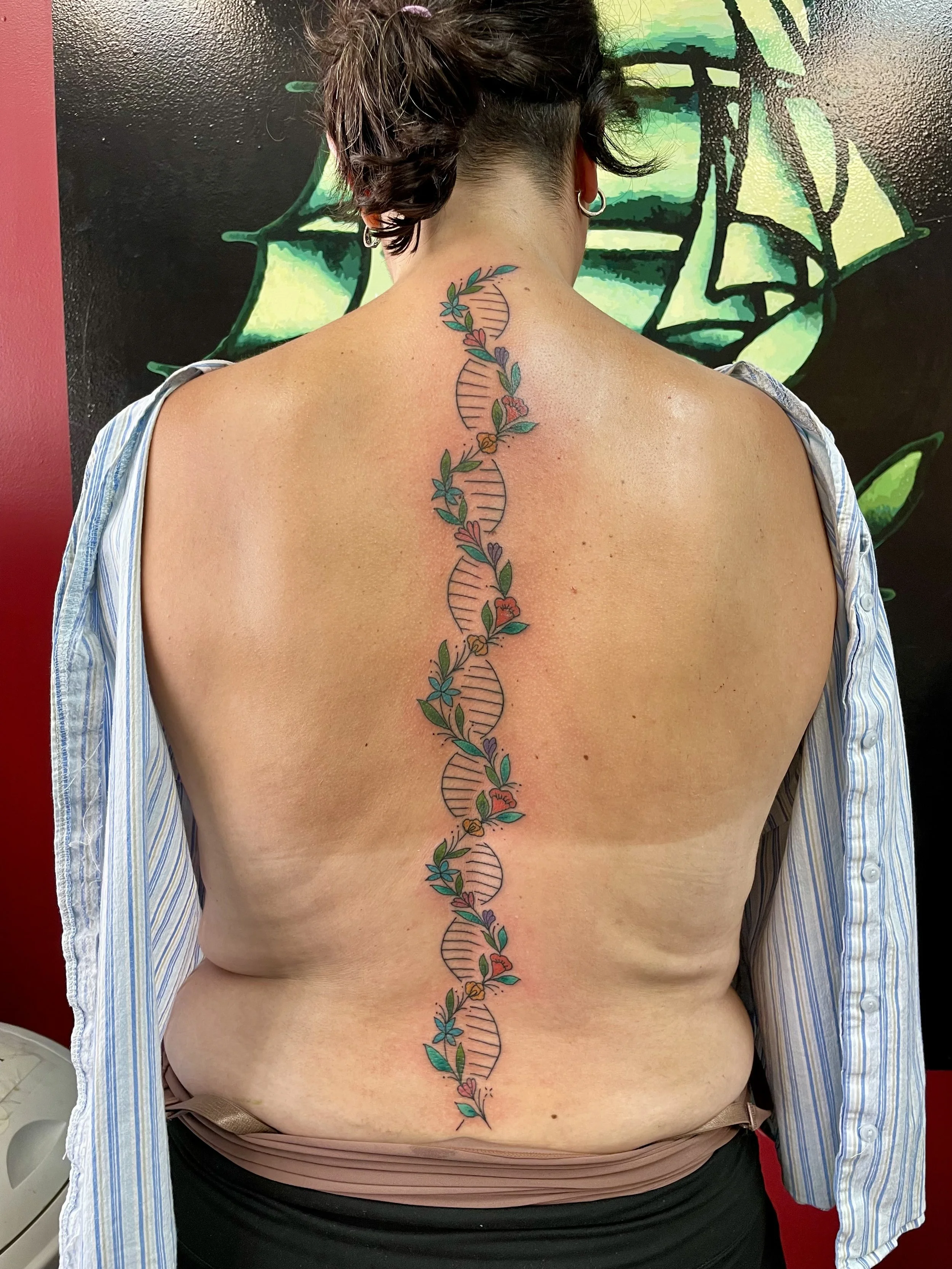 Fine line color spine tattoo featuring a full-length DNA double helix running from the neck to the lower back, intertwined with delicate botanical illustrations of red, orange, and purple wildflowers with green leaves throughout, custom tattoo by Nic