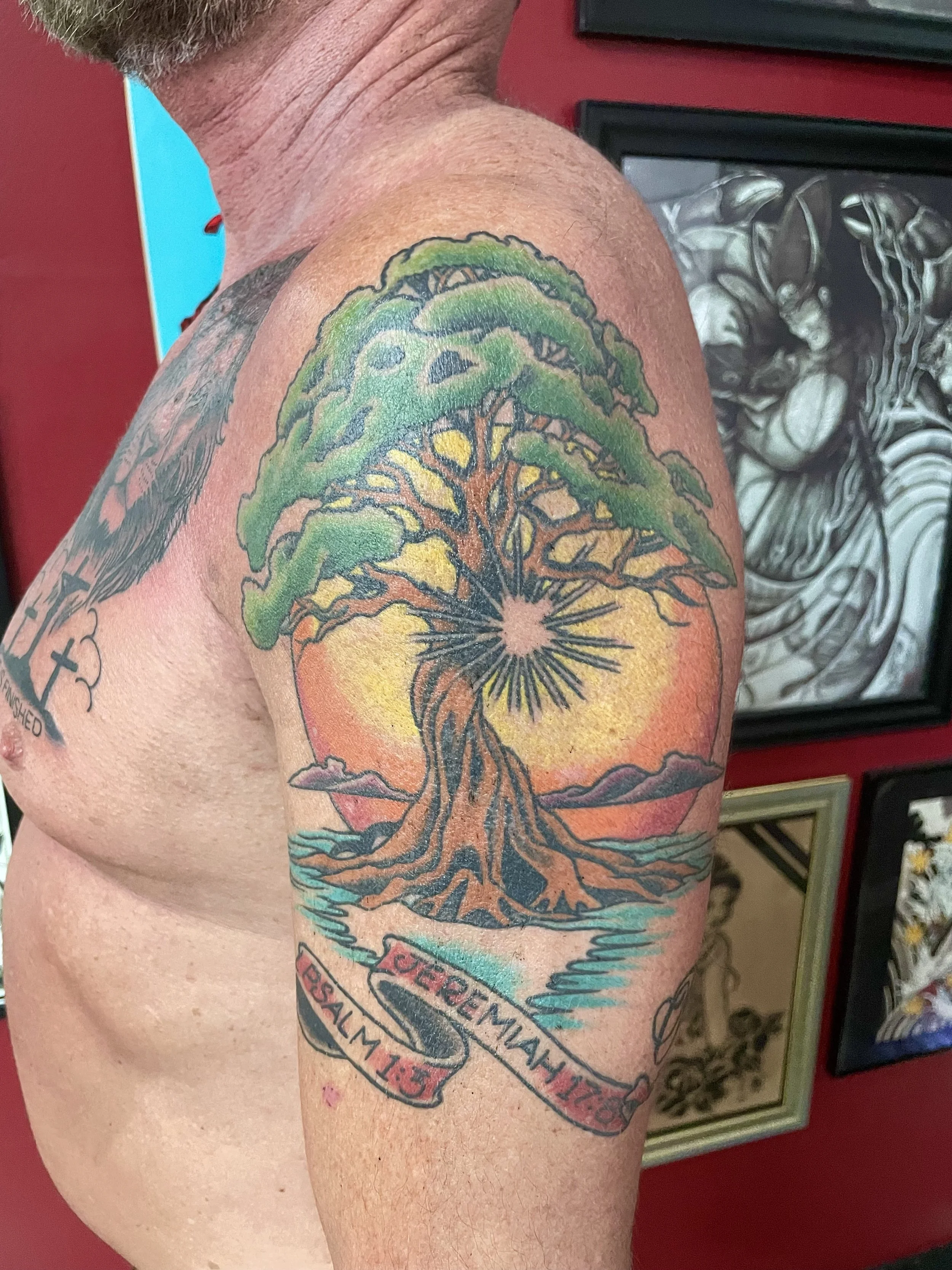 Traditional color upper arm tattoo featuring a sprawling Tree of Life with a lush green canopy and twisted exposed roots, set against a warm golden and orange sunset, with teal water below and two scripture scroll banners reading "Jeremiah 17:8" and 