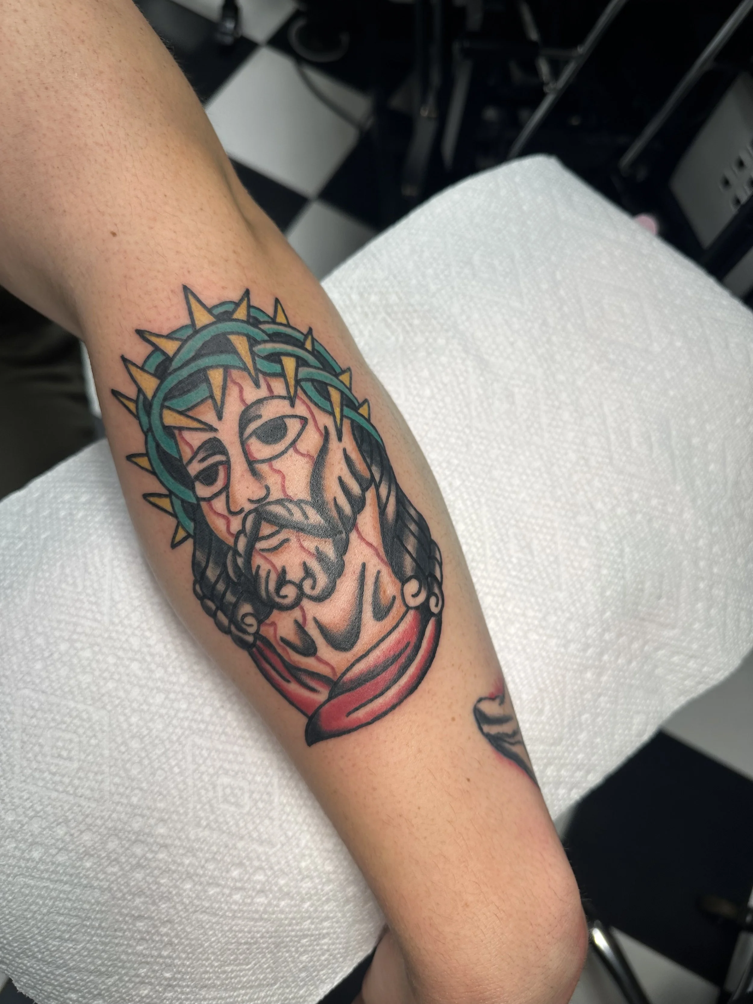 Traditional American color forearm tattoo featuring a bold portrait of Jesus wearing a teal crown of thorns with gold spikes, rendered with strong linework and a red banner beneath, custom tattoo by Dan at Big Kahuna Tattoo in Boca Raton, FL.