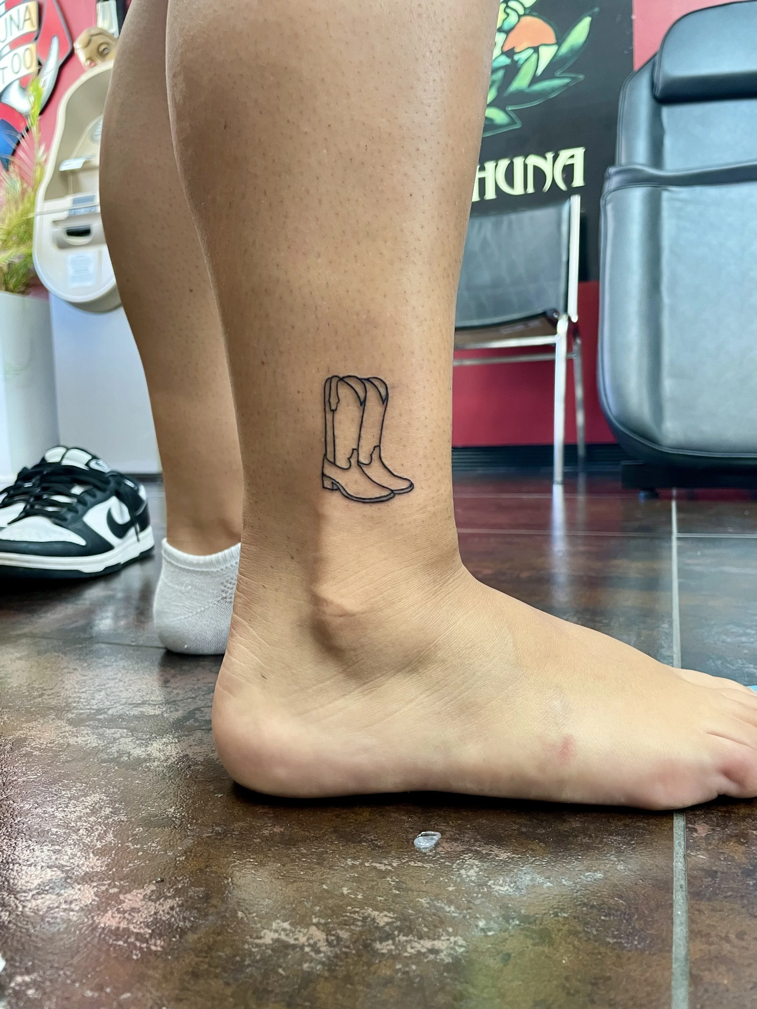 Minimalist fine line ankle tattoo featuring a pair of cowboy boots in a simple clean outline style, custom tattoo by Nick at Big Kahuna Tattoo in Boca Raton, FL.