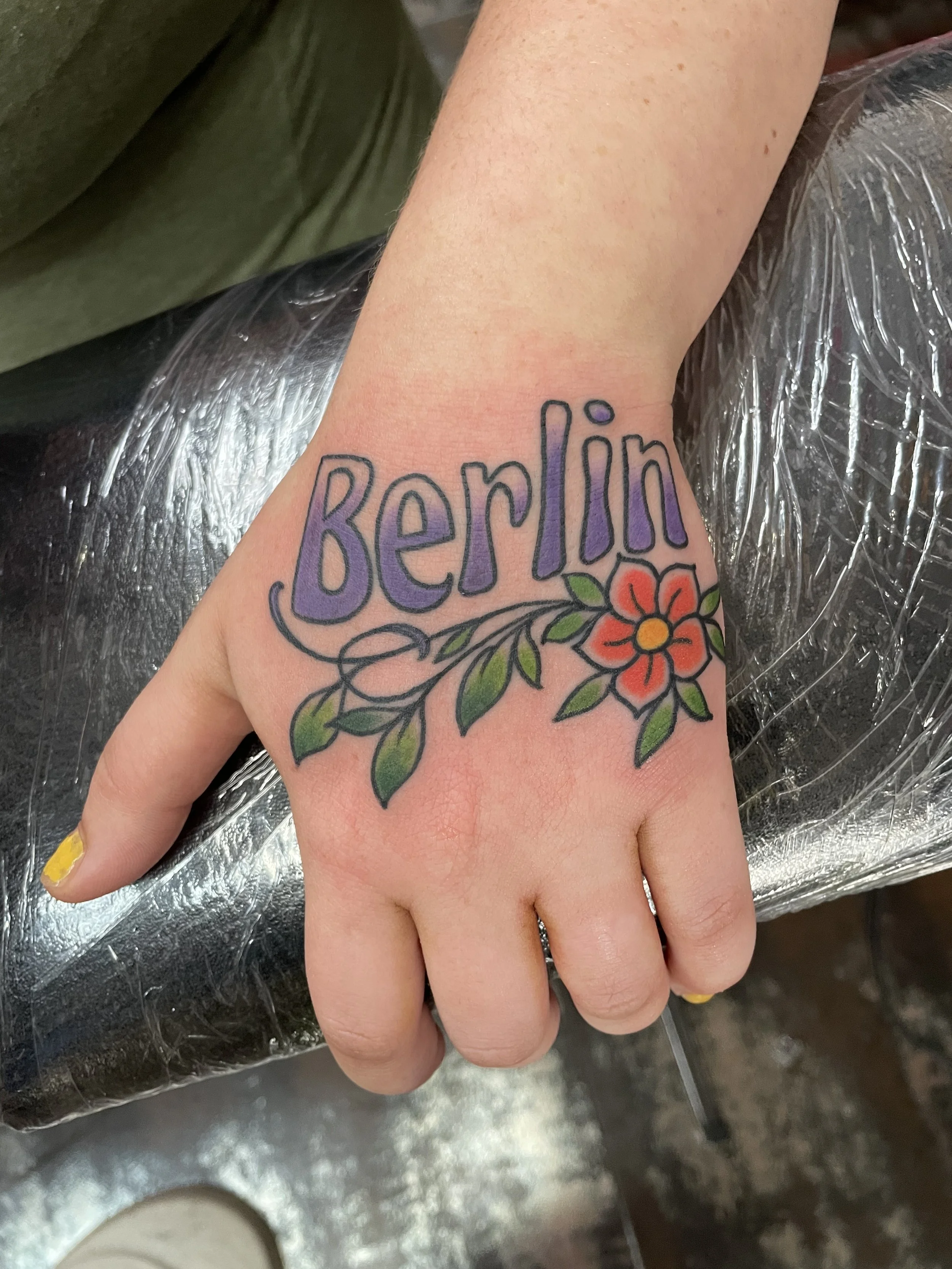 Color hand tattoo featuring bold purple block lettering spelling "Berlin" across the back of the hand, accented below with a red flower and green leafy stem, custom tattoo by Nick at Big Kahuna Tattoo in Boca Raton, FL.