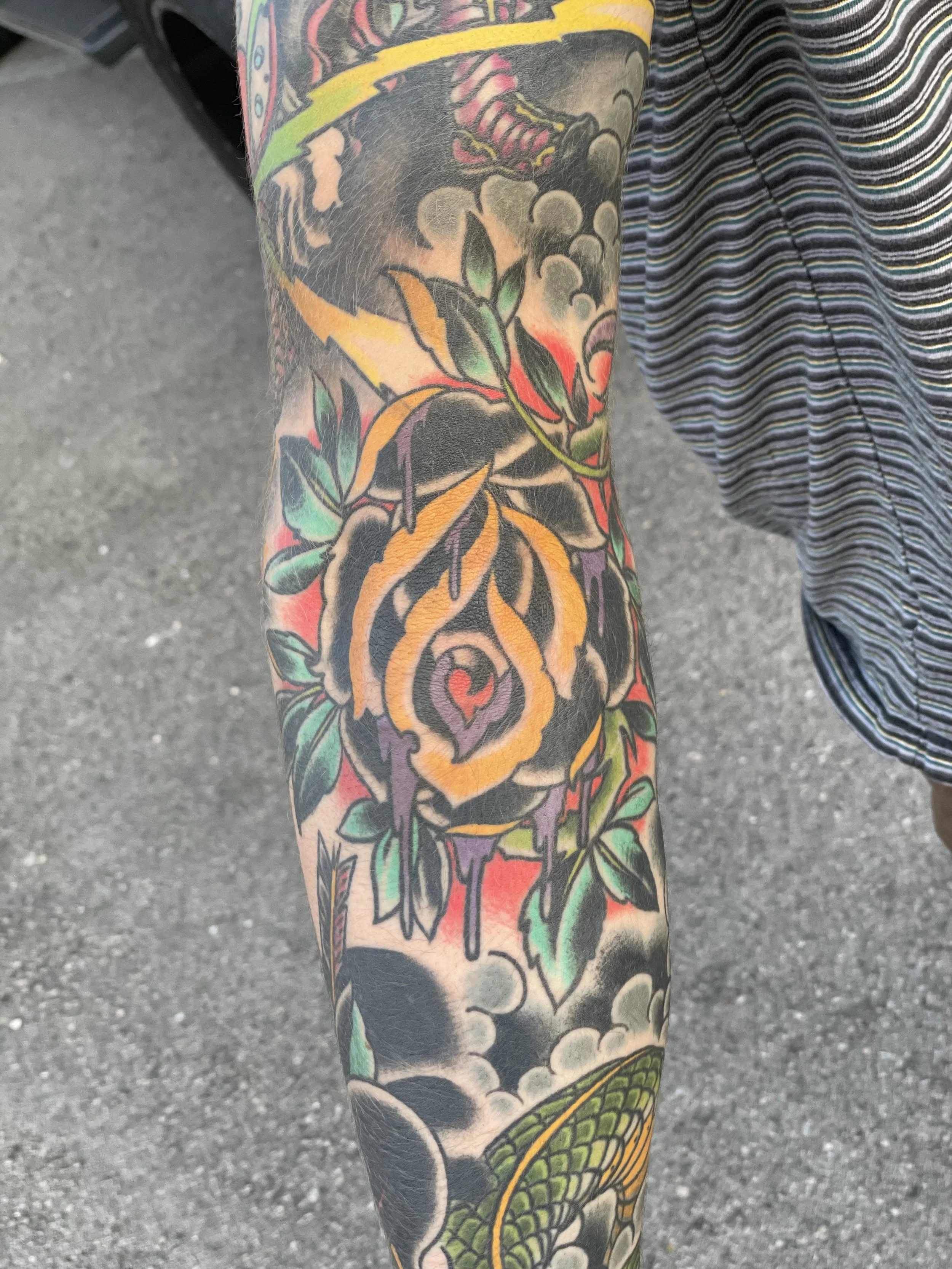 Traditional color forearm sleeve tattoo featuring a large dark rose with gold-outlined petals, set among dark foliage, red accents, clouds, and green snake scales as part of a larger sleeve composition, custom tattoo by Nick at Big Kahuna Tattoo in B