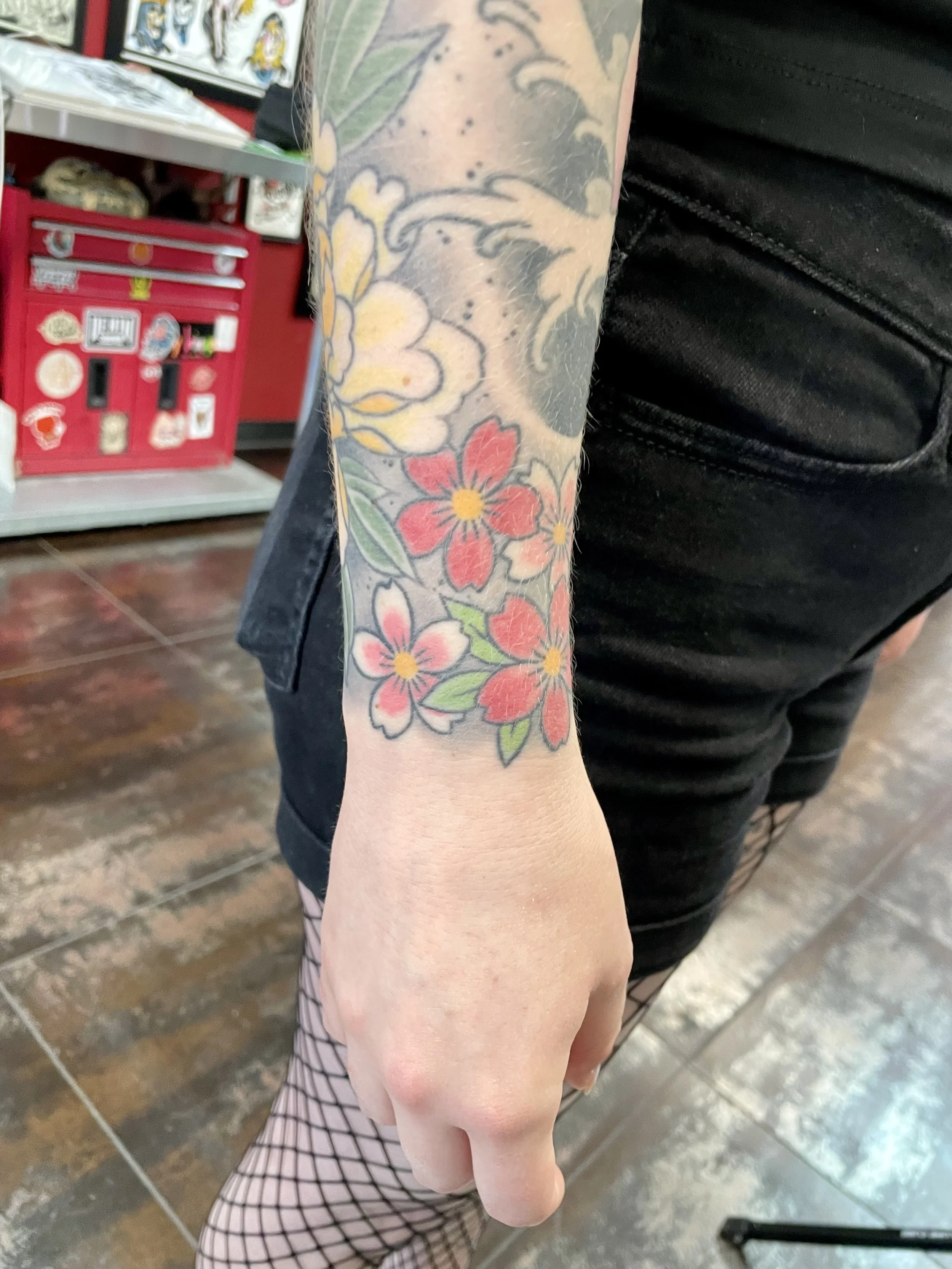 A cluster of red and pink cherry blossoms with golden centers at the wrist, connecting up to a soft yellow peony above. Carefully composed addition to an existing Japanese sleeve. Color wrist tattoo by Nick at Big Kahuna Tattoo in Boca Raton, FL.