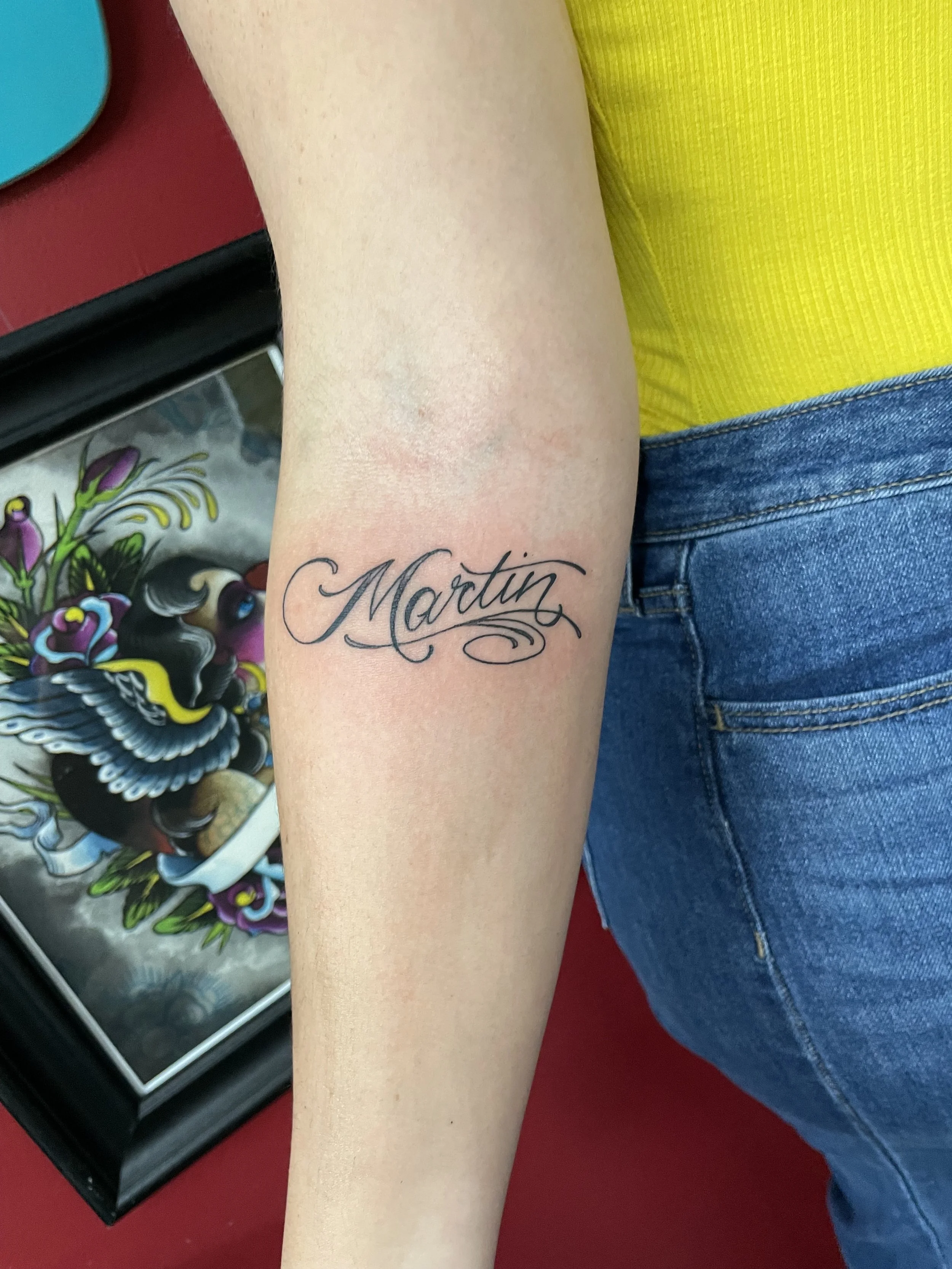 Fine line forearm tattoo featuring the name "Martin" in flowing ornate cursive lettering with elegant tail flourishes, custom tattoo by Nick at Big Kahuna Tattoo in Boca Raton, FL.