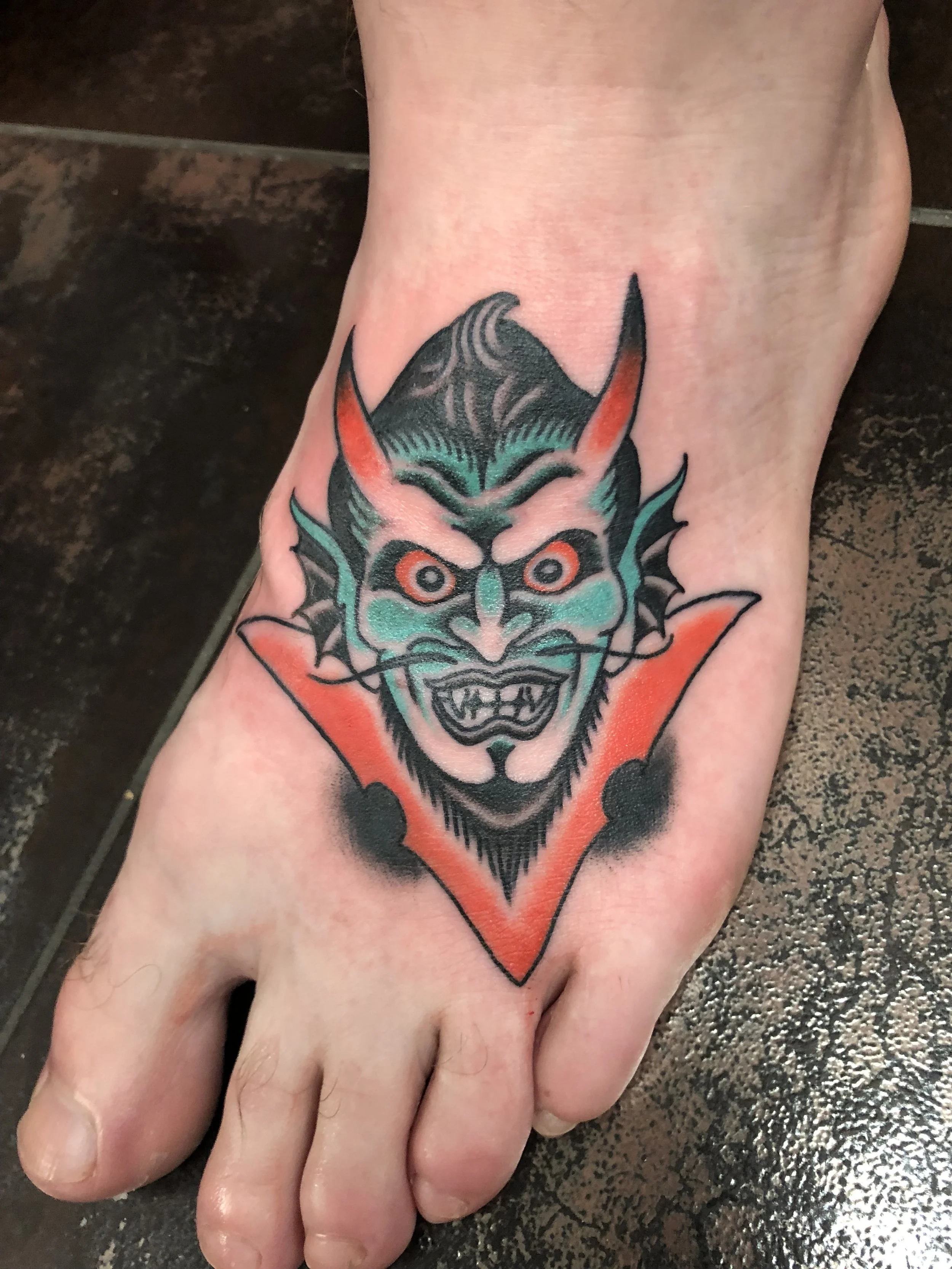 Traditional American color foot tattoo featuring a grinning teal devil face with red horns inside a bold red triangle, custom tattoo by Nick at Big Kahuna Tattoo in Boca Raton, FL.