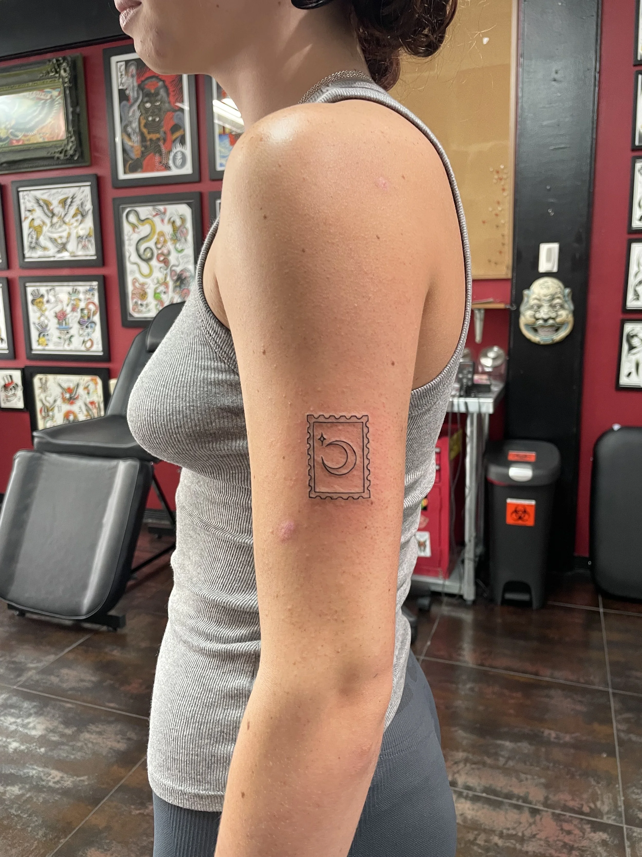Fine line shoulder tattoo featuring a minimalist postage stamp outline containing a crescent moon and a four-pointed star, custom tattoo by Nick at Big Kahuna Tattoo in Boca Raton, FL.