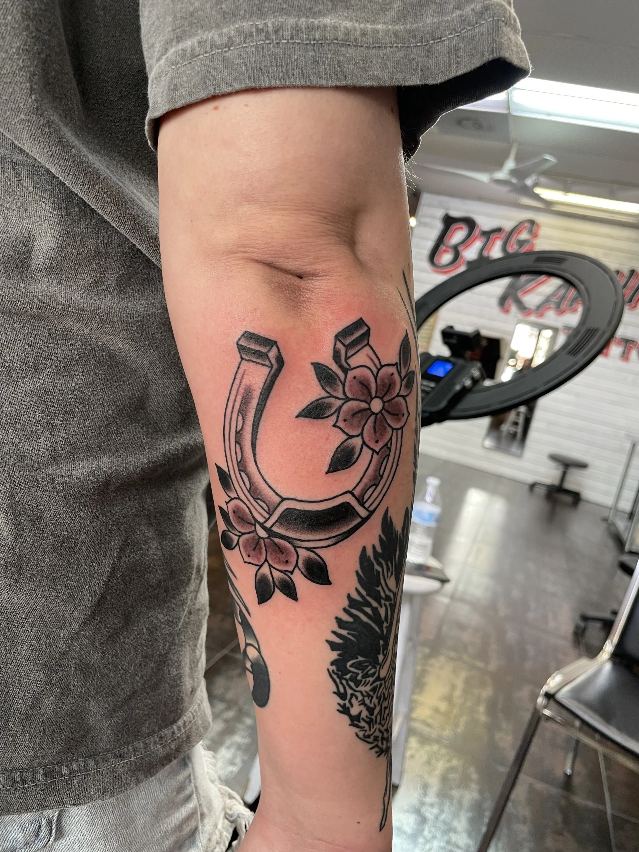 Black and grey forearm tattoo featuring a classic horseshoe wrapped with soft blossoms and dark leaves for luck and beauty, set alongside existing sleeve work, custom tattoo by Nick at Big Kahuna Tattoo in Boca Raton, FL.