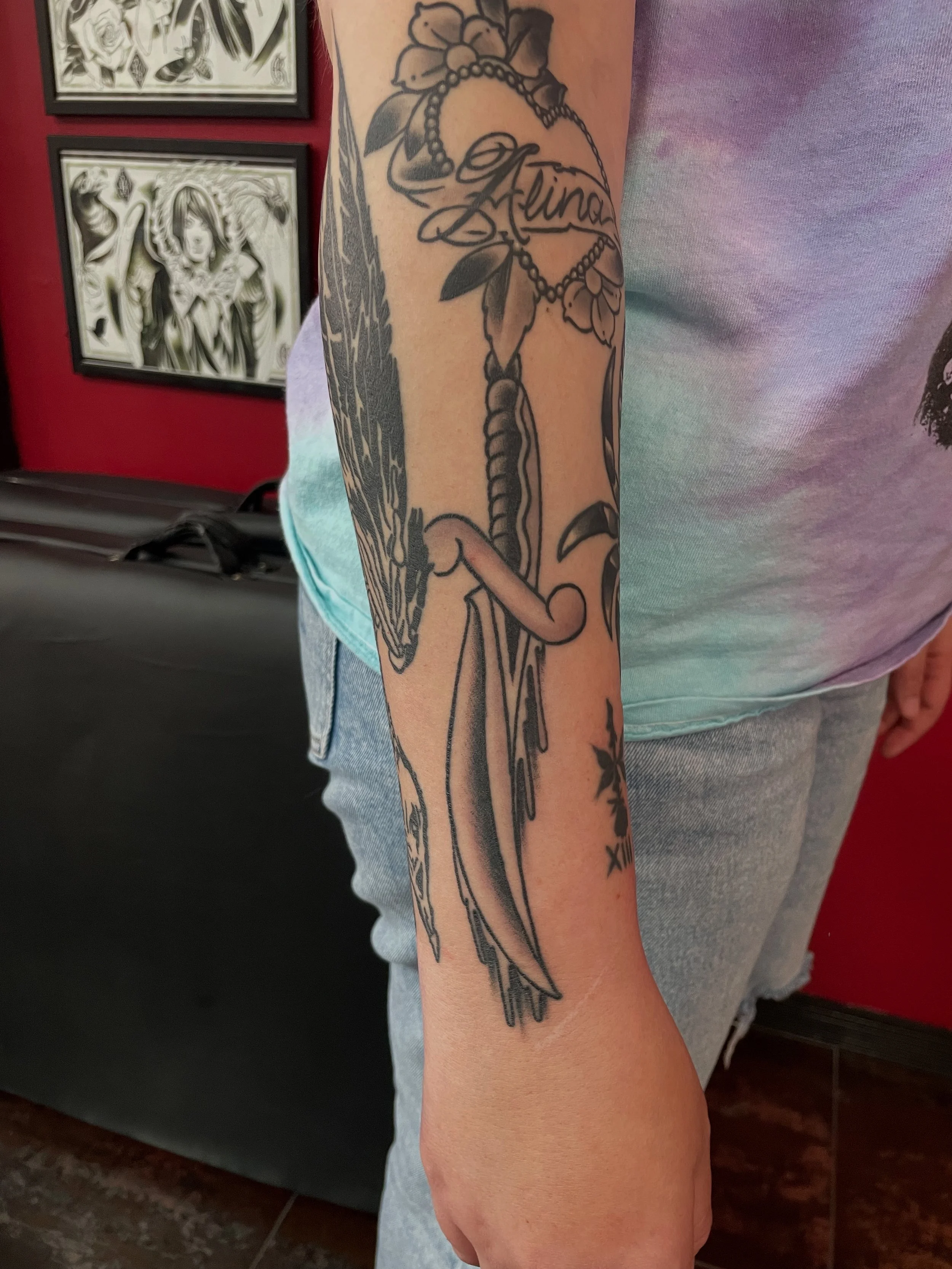 Black and grey forearm sleeve tattoo featuring a long dripping dagger with a wrapped handle, set among existing sleeve work including a floral heart banner, custom tattoo by Nick at Big Kahuna Tattoo in Boca Raton, FL.