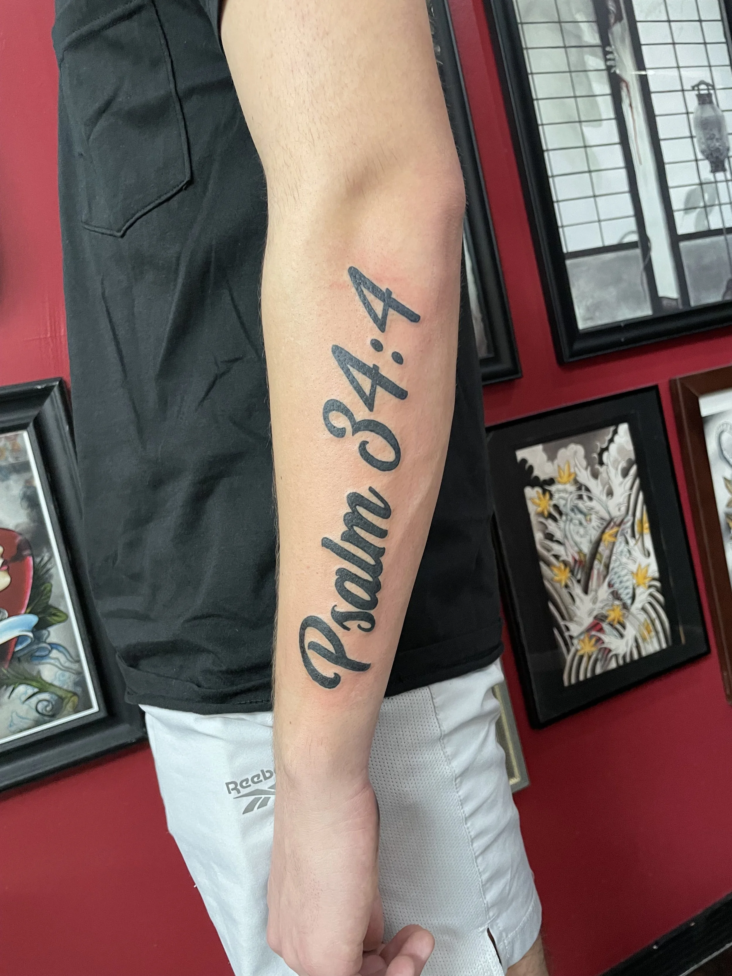 Bold black script forearm tattoo reading "Psalm 34:4" in large flowing cursive lettering running diagonally along the length of the forearm, custom tattoo by Nick at Big Kahuna Tattoo in Boca Raton, FL.