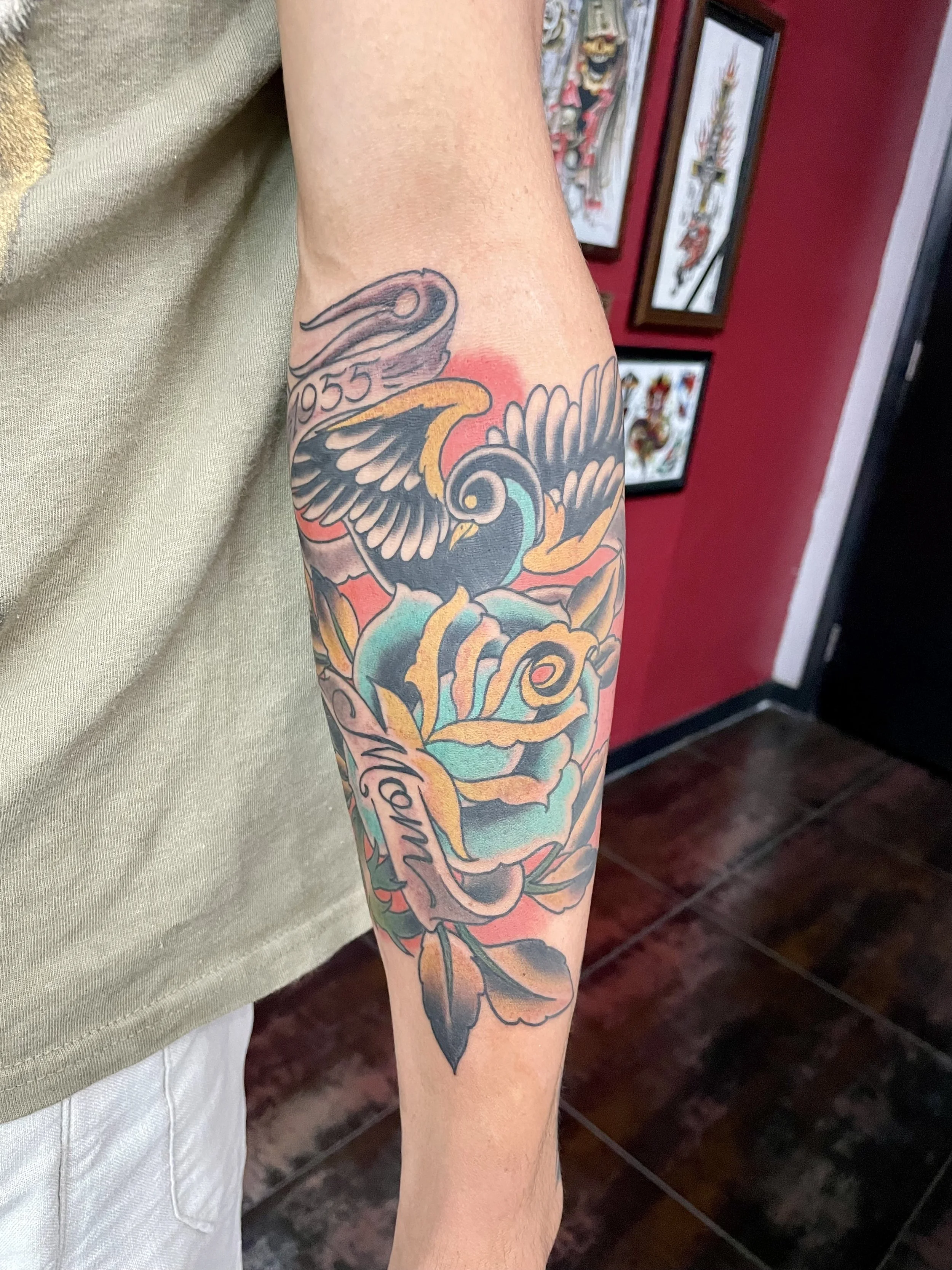 Traditional American color forearm tattoo featuring a bold black swallow above a large teal and gold rose, with two scroll banners reading "1955" and "Mom," custom tattoo by Nick at Big Kahuna Tattoo in Boca Raton, FL.