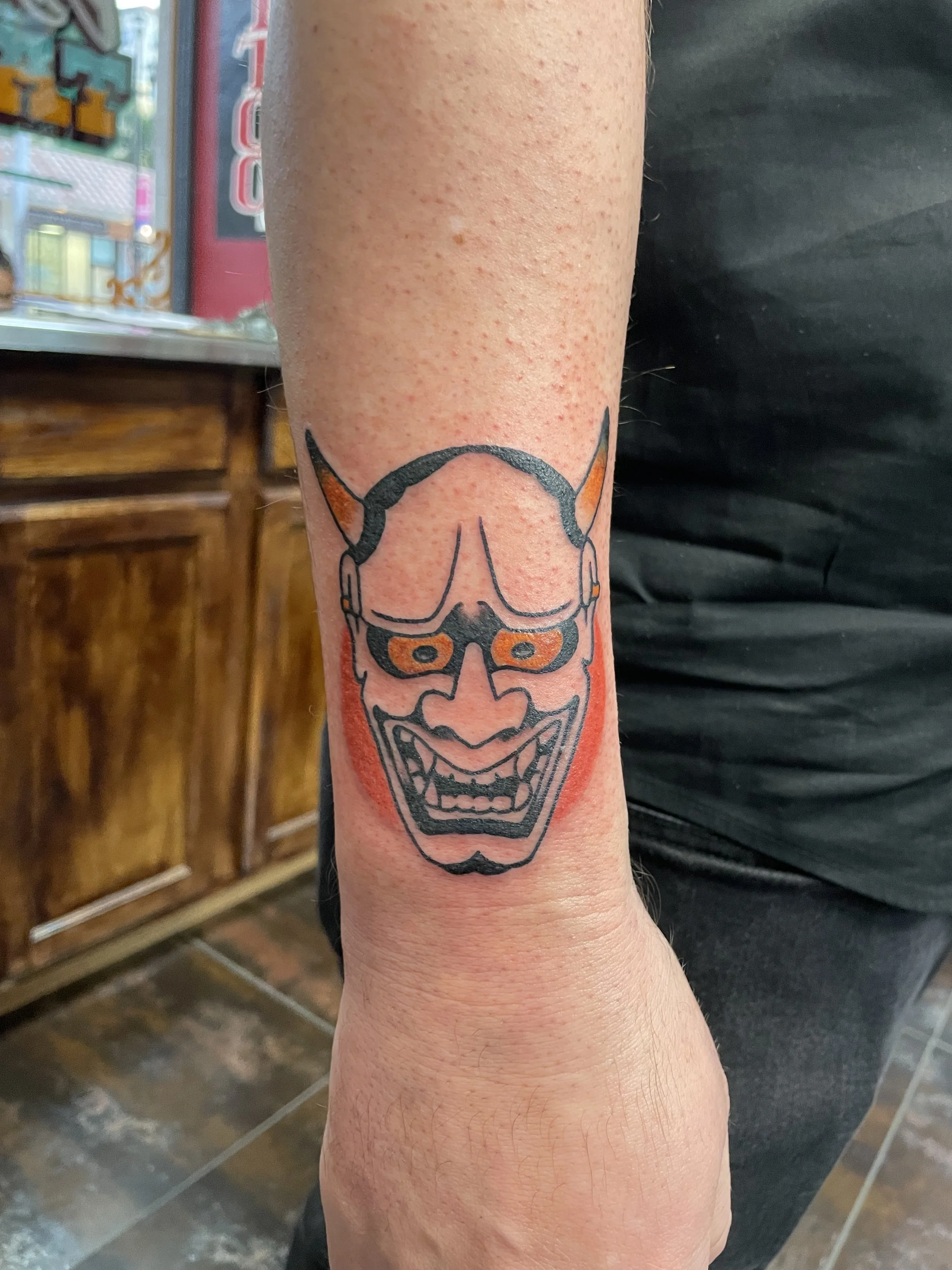 Small but impossible to ignore. A bold Hannya mask with orange horns and eye sockets, set against a soft red background shadow on the wrist. Japanese wrist tattoo by Nick at Big Kahuna Tattoo in Boca Raton, FL.