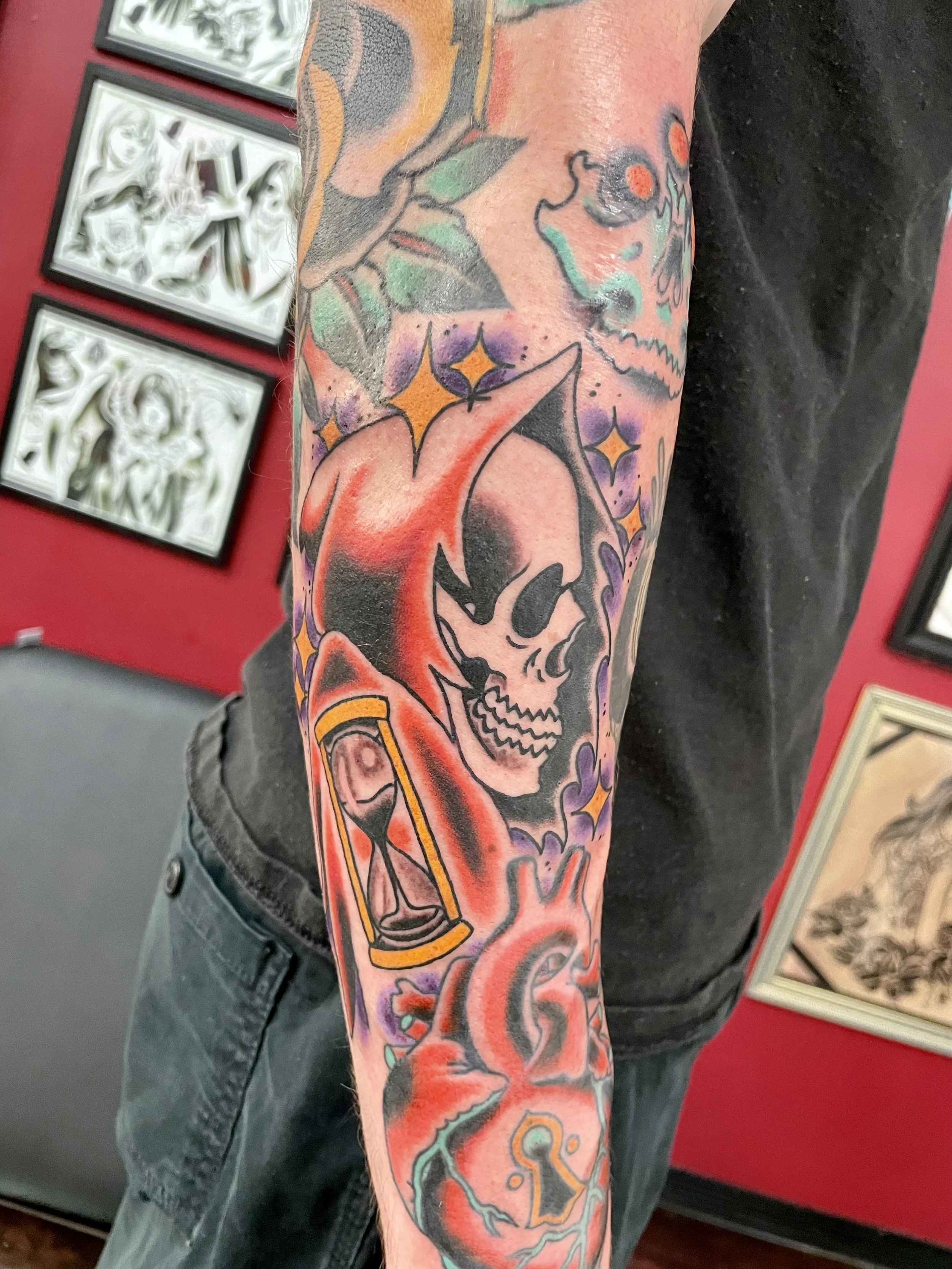 Traditional color forearm sleeve tattoo featuring a stylized Grim Reaper skull in a red shroud surrounded by gold four-pointed stars, with a gold hourglass at his side, set among existing sleeve work, custom tattoo by Nick at Big Kahuna Tattoo in Boc