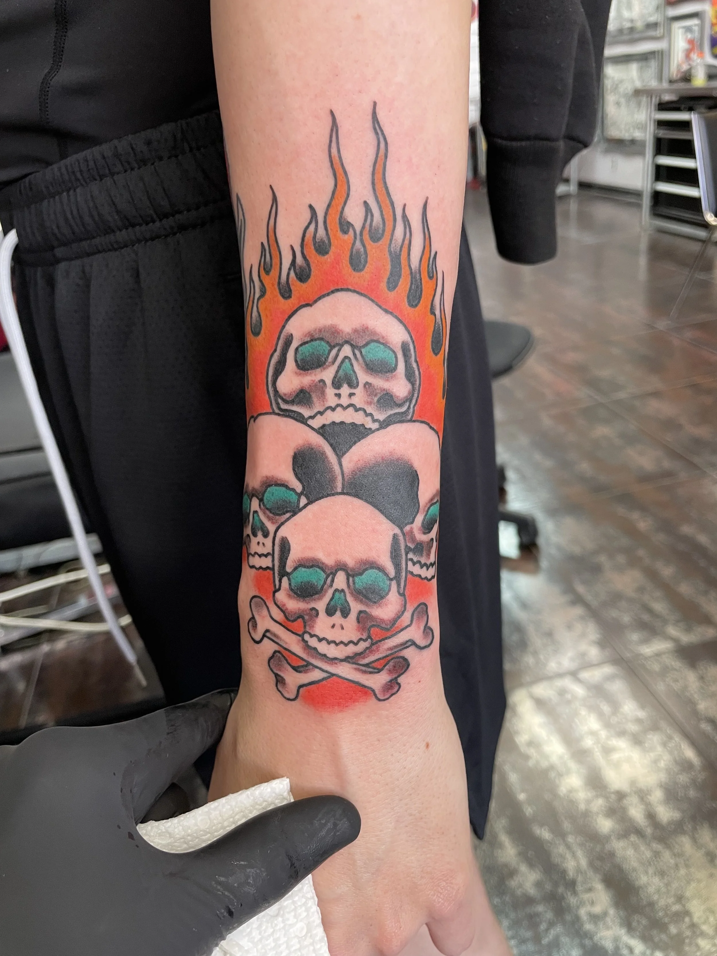 Traditional American color forearm tattoo featuring a stacked trio of skulls with teal eye sockets rising through bold orange and red flames, with crossed bones at the base, custom tattoo by Nick at Big Kahuna Tattoo in Boca Raton, FL.