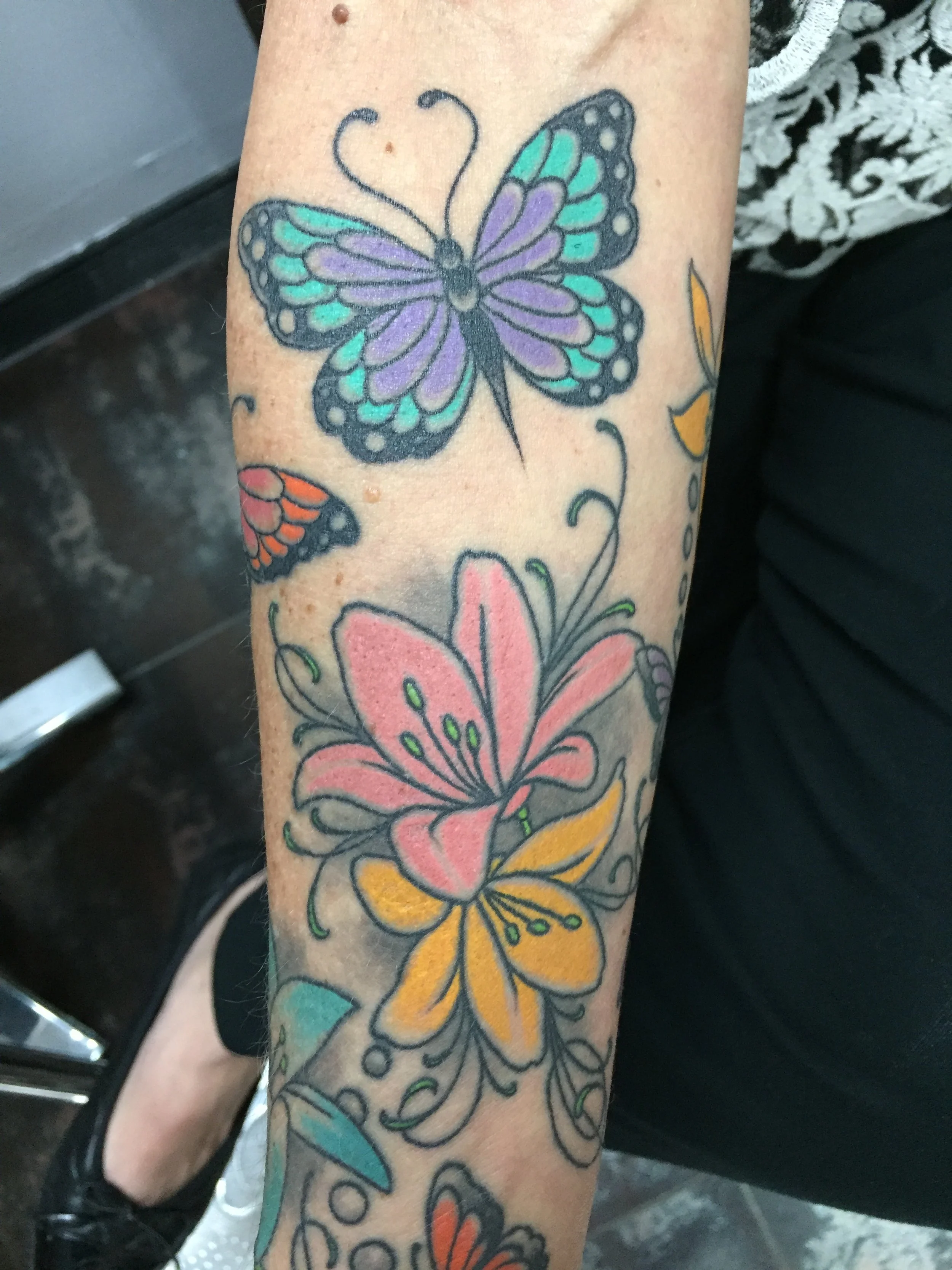 Colorful traditional forearm tattoo featuring a large purple and teal butterfly alongside pink and yellow lilies with decorative vines, custom tattoo by Nick at Big Kahuna Tattoo in Boca Raton, FL.
