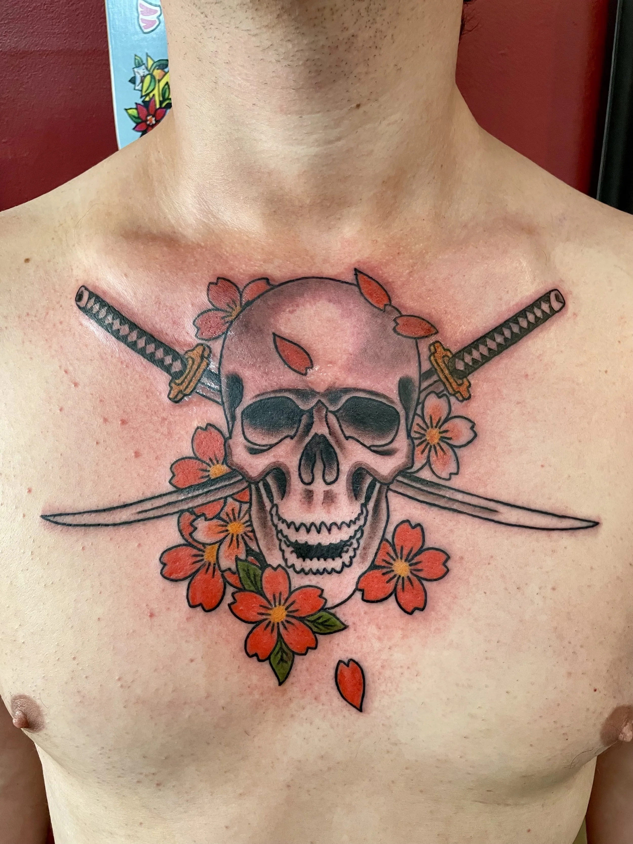 Beauty and violence in one composition. A bold skull centered over two crossed katanas with detailed wrapped handles, surrounded by a ring of vivid red cherry blossoms scattering across the chest. Japanese color chest tattoo by Nick at Big Kahuna Tat