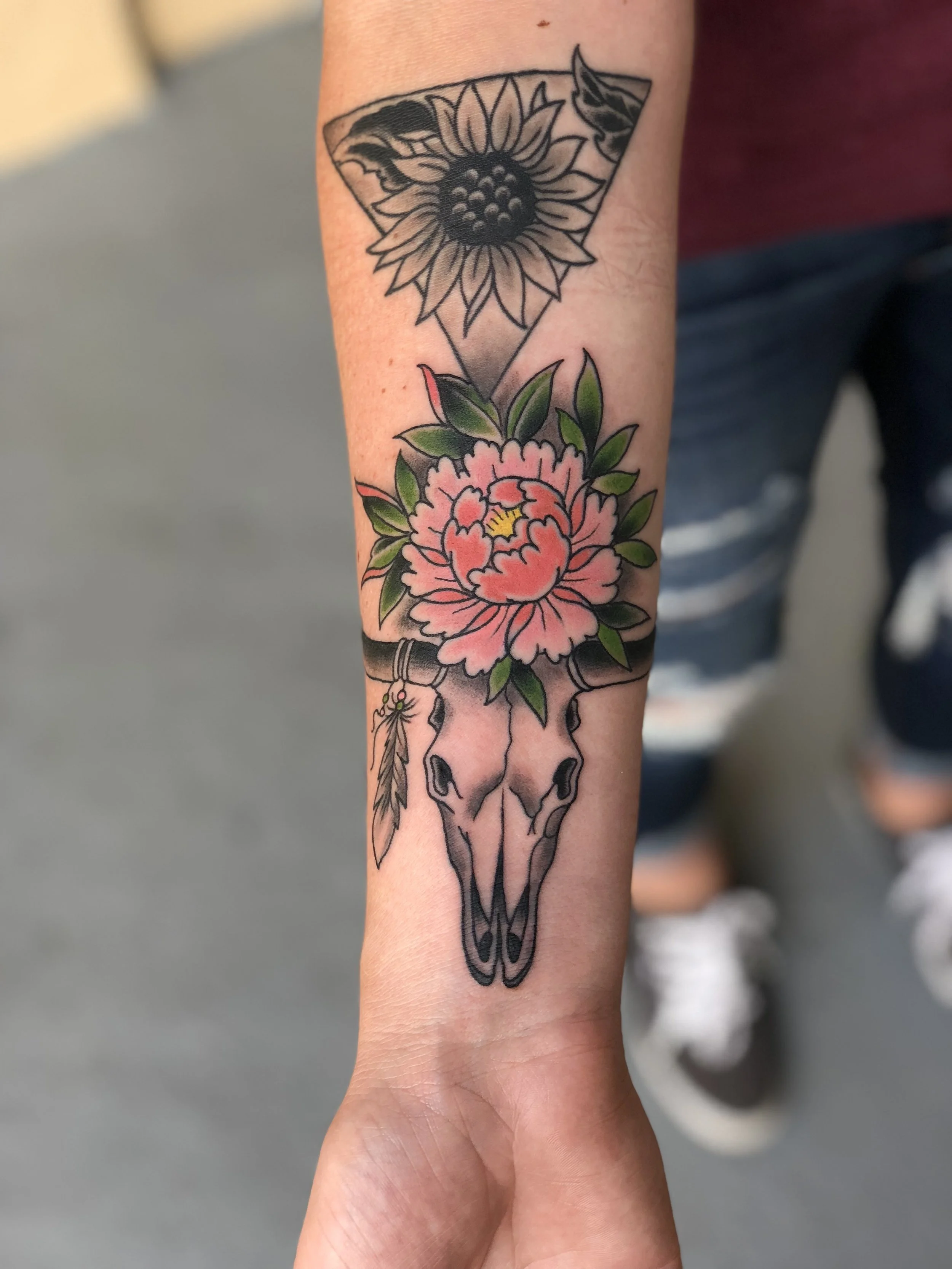 Neo traditional color forearm tattoo featuring a longhorn bull skull topped with a pink peony and green leaves, with a hanging feather and a black and grey sunflower in a triangle above, custom tattoo by Nick at Big Kahuna Tattoo in Boca Raton, FL.