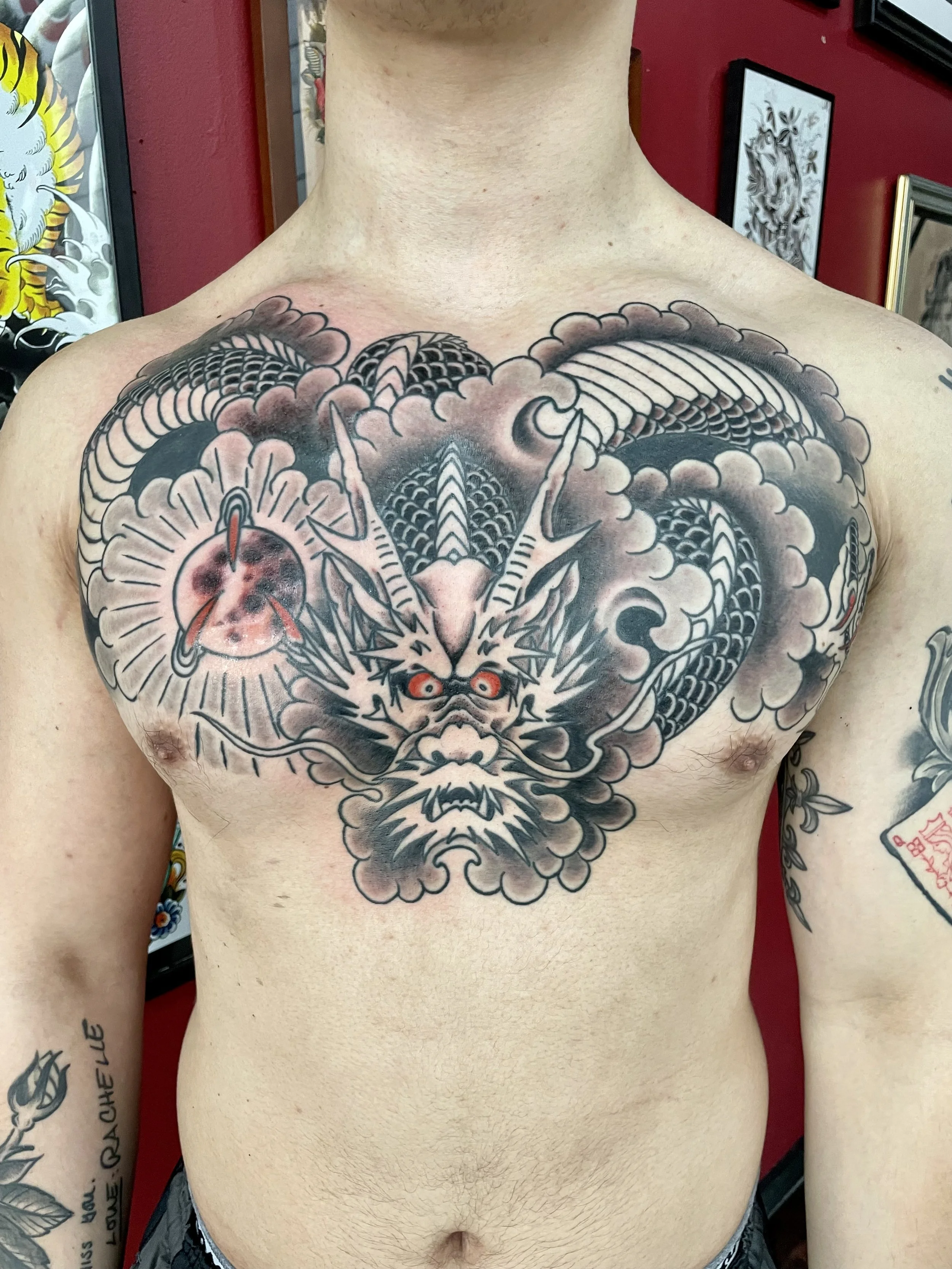 Large Japanese black and grey chest piece tattoo featuring a commanding dragon with piercing red eyes, sweeping horns, and detailed scales, its serpentine body coiling symmetrically across the full chest surrounded by dramatic clouds and a glowing or