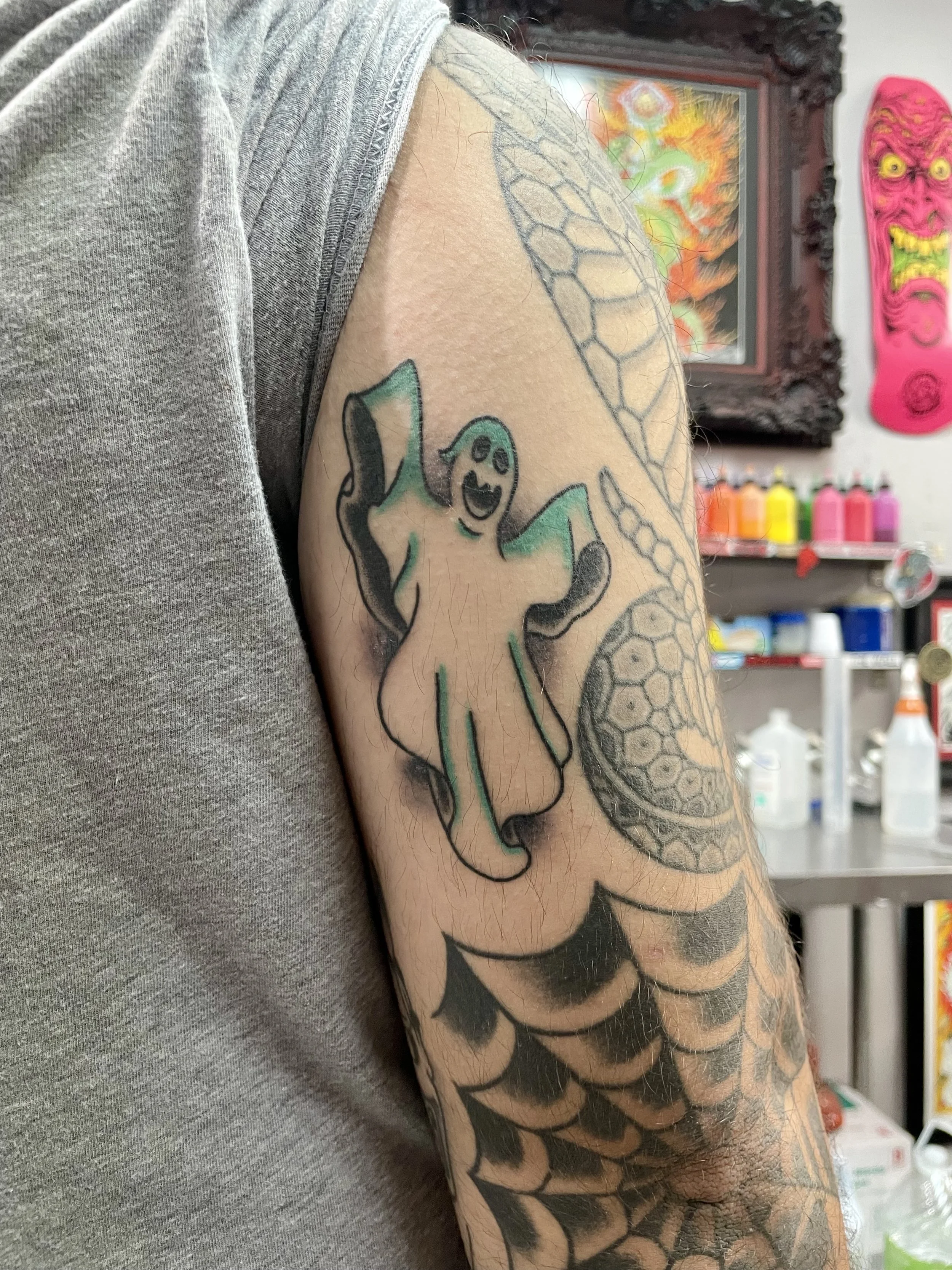 Traditional American upper arm tattoo featuring a friendly floating ghost in teal and white with a smiling face and raised arms, set against a dark shadow background alongside existing spiderweb sleeve work, custom tattoo by Nick at Big Kahuna Tattoo