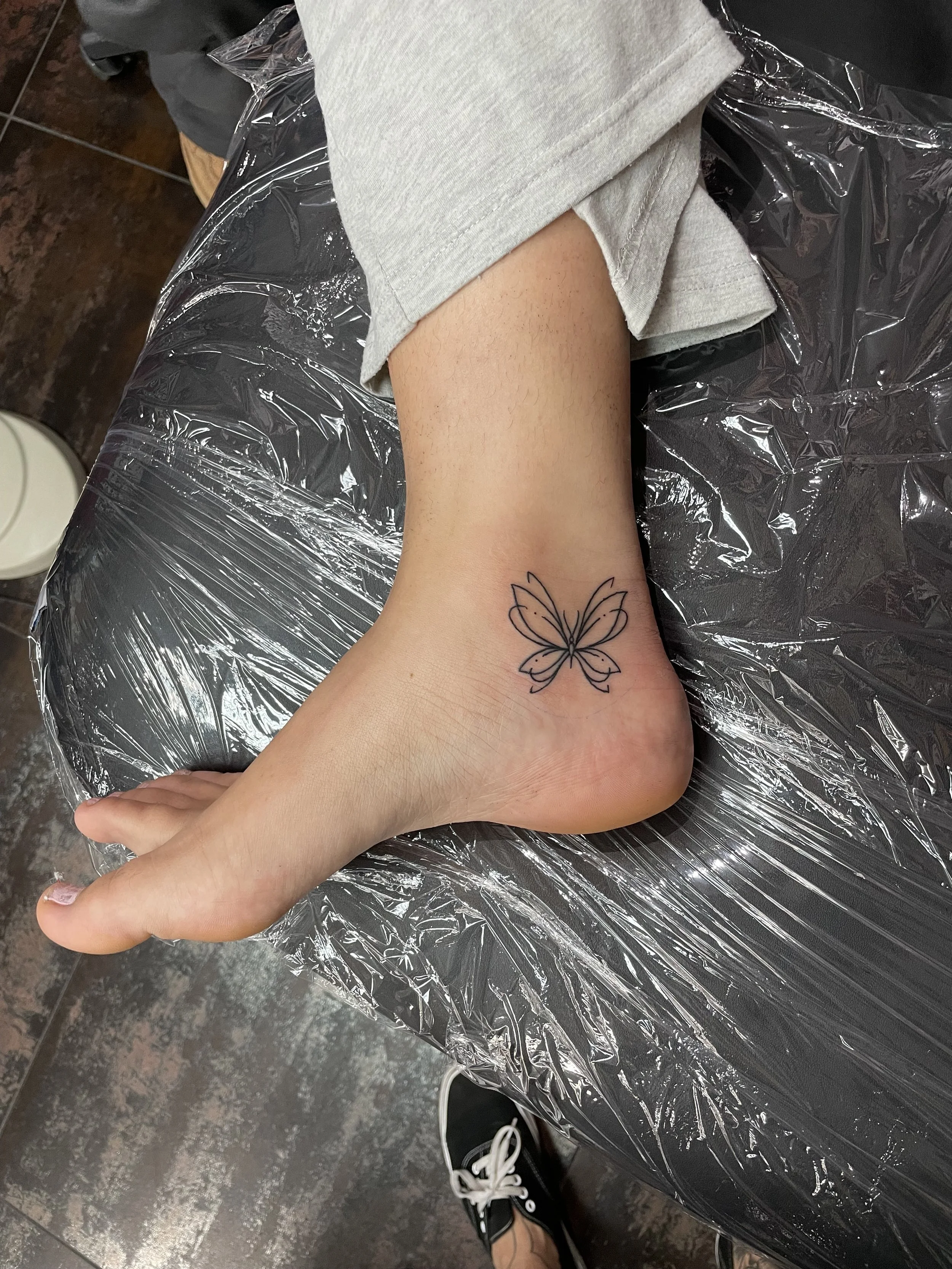 Fine line ankle tattoo featuring a delicate minimalist butterfly outline with softly rounded wings, custom tattoo by Nick at Big Kahuna Tattoo in Boca Raton, FL.
