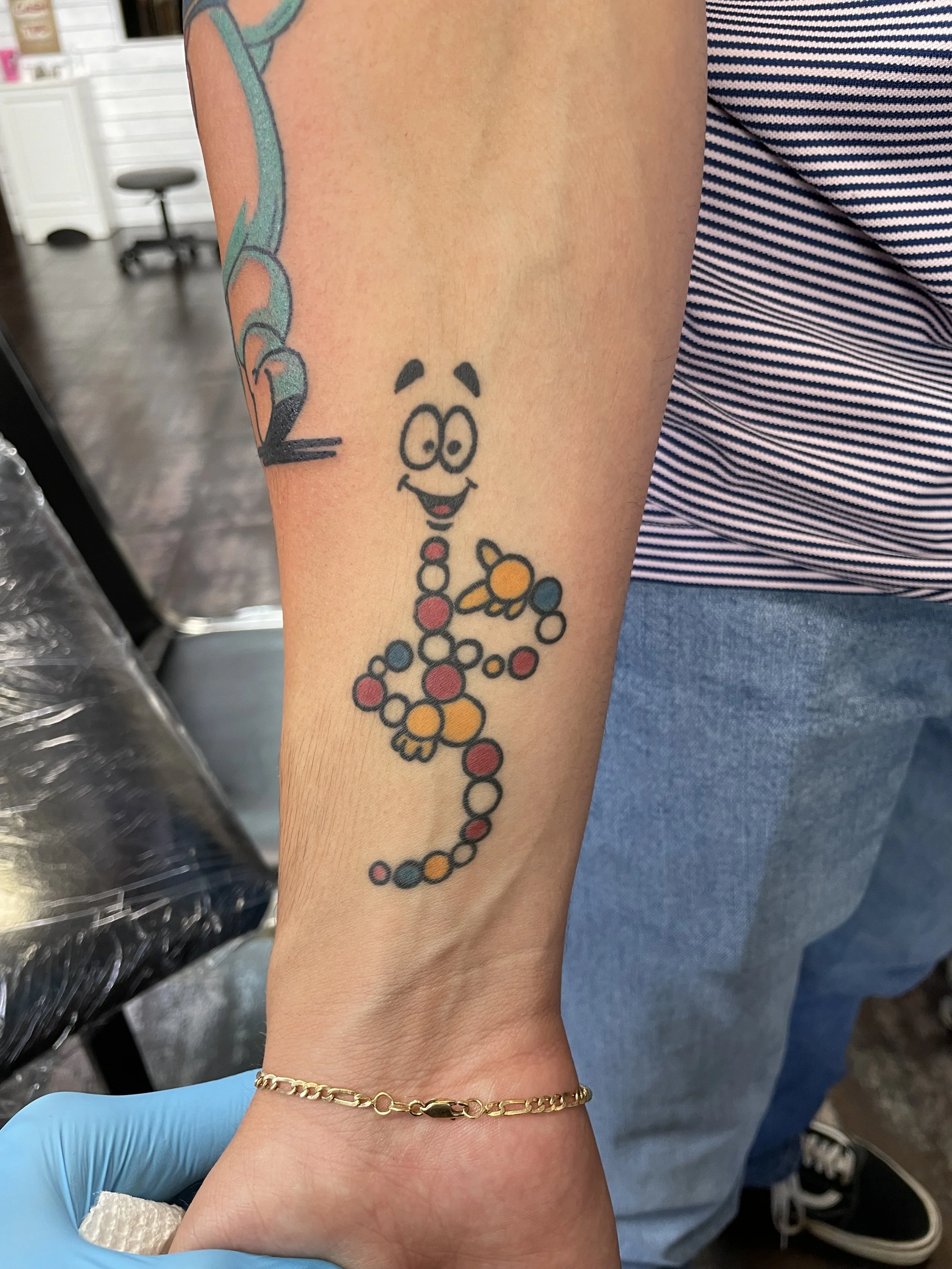 Colorful cartoon forearm tattoo featuring Mr. DNA from Jurassic Park, rendered as a cheerful animated DNA strand figure with red, yellow, teal, and white molecular dots, custom tattoo by Nick at Big Kahuna Tattoo in Boca Raton, FL.