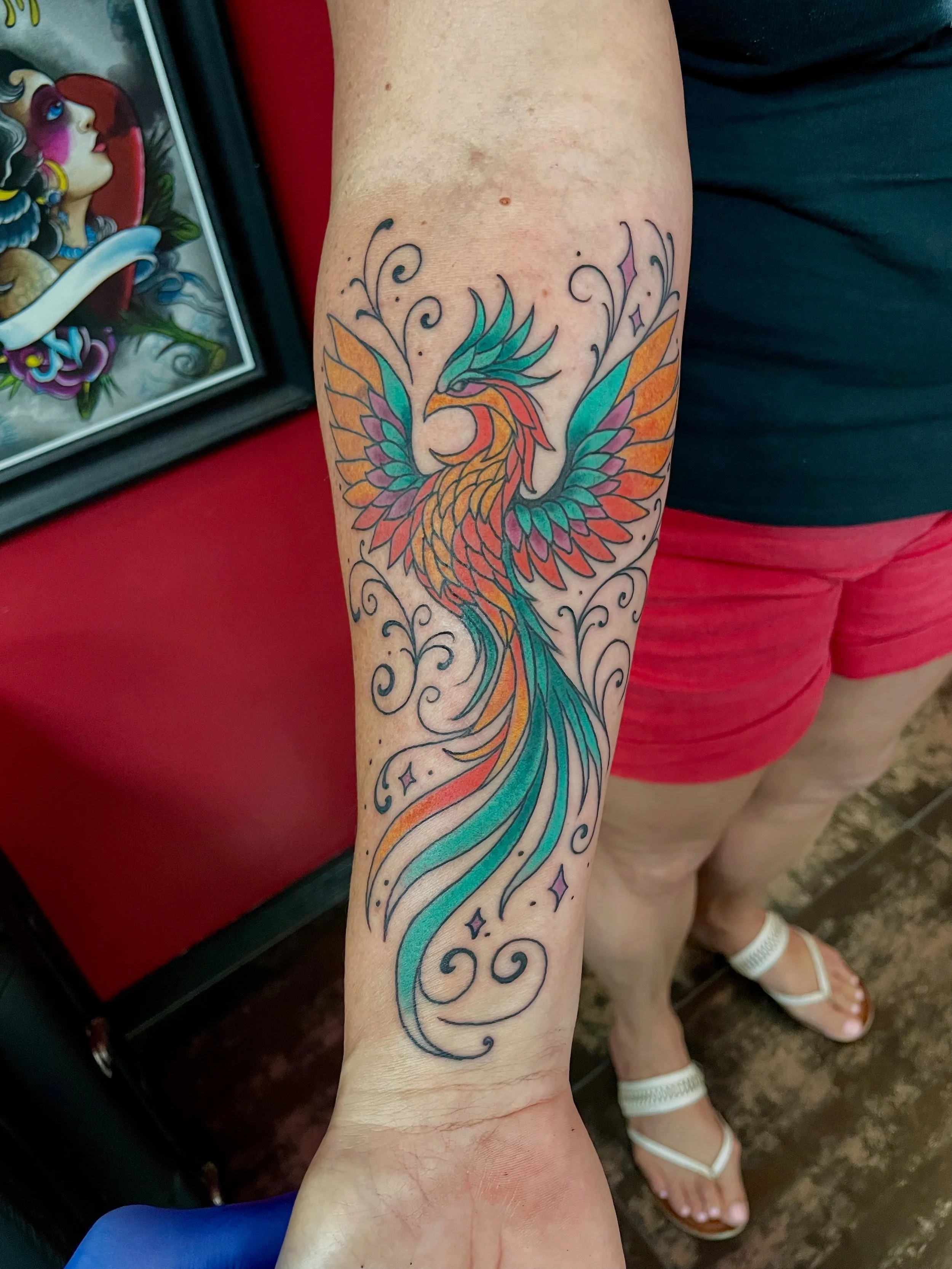 Fine line color forearm tattoo featuring a vibrant rising phoenix with outstretched wings in teal, orange, red, and pink, its long tail feathers swirling into elegant decorative flourishes and four-pointed stars down the forearm, custom tattoo by Nic