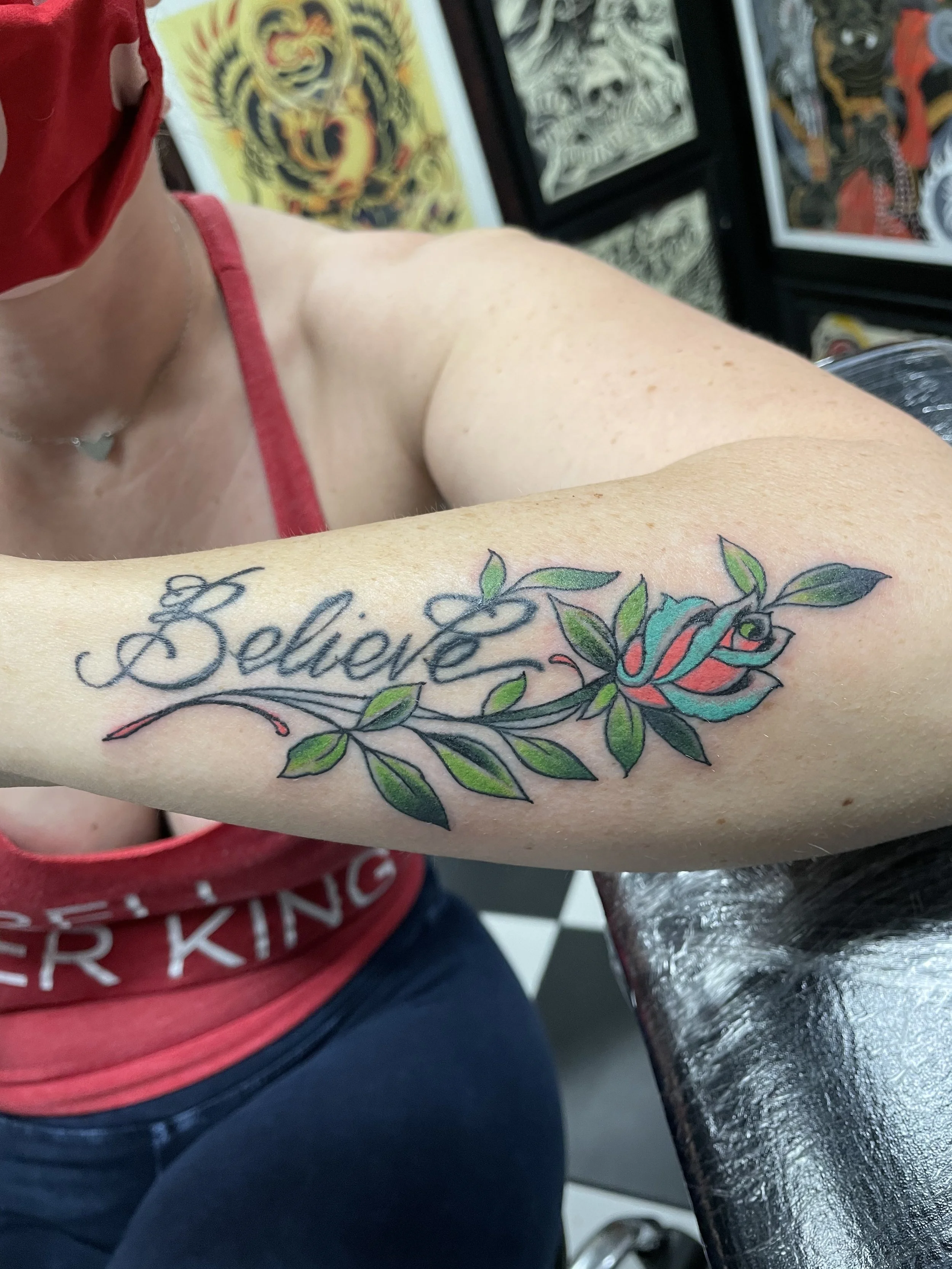 Color forearm tattoo featuring elegant script lettering reading "Believe" woven through a flowering rose branch with green leaves and a teal and coral rose bloom, custom tattoo by Nick at Big Kahuna Tattoo in Boca Raton, FL.