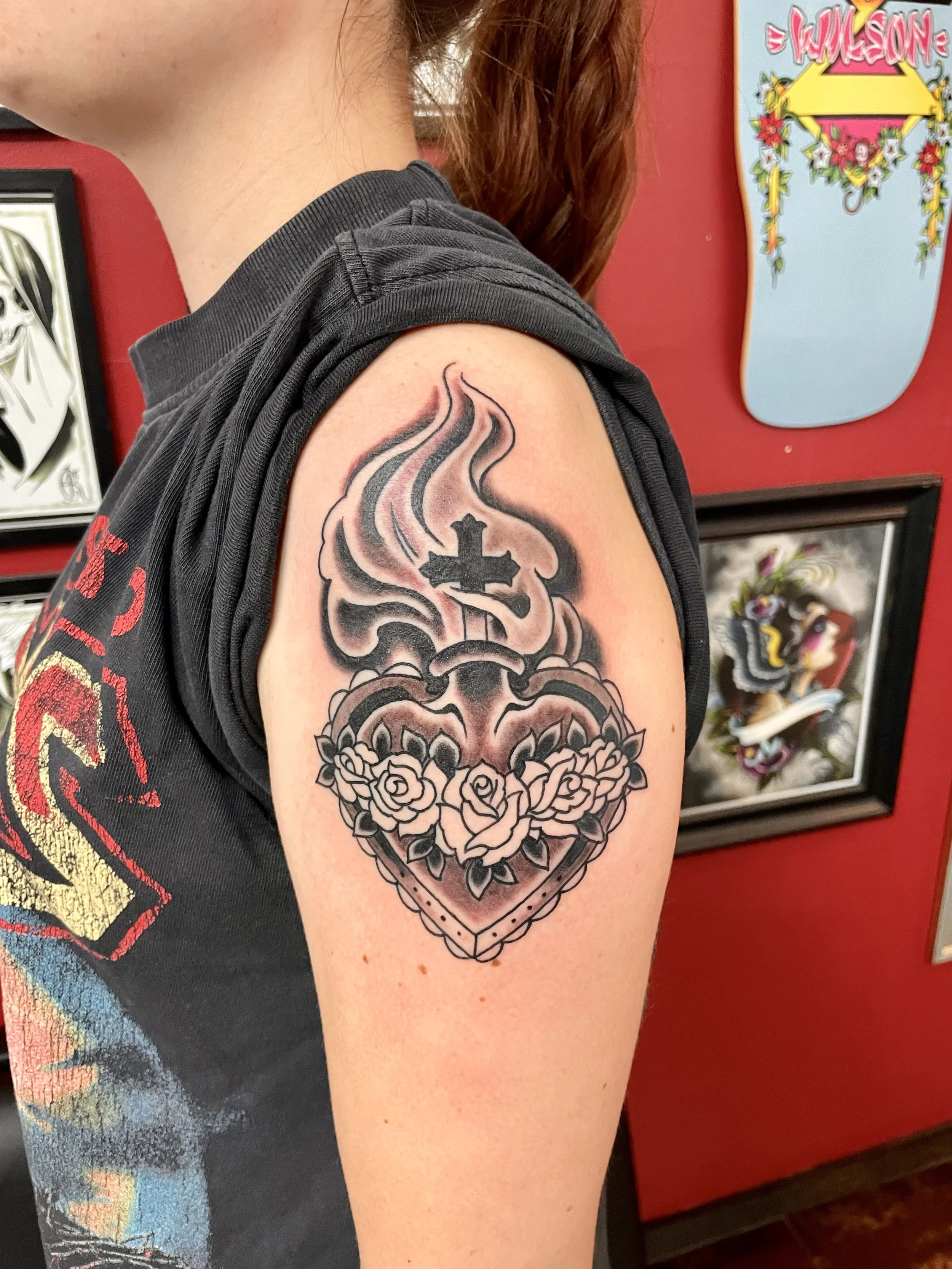 A timeless religious image rendered with conviction. A sacred heart crowned by a flaming cross and framed by three blooming roses across the base, the whole piece glowing with soft shading against deep black. Black and grey shoulder tattoo by Nick at
