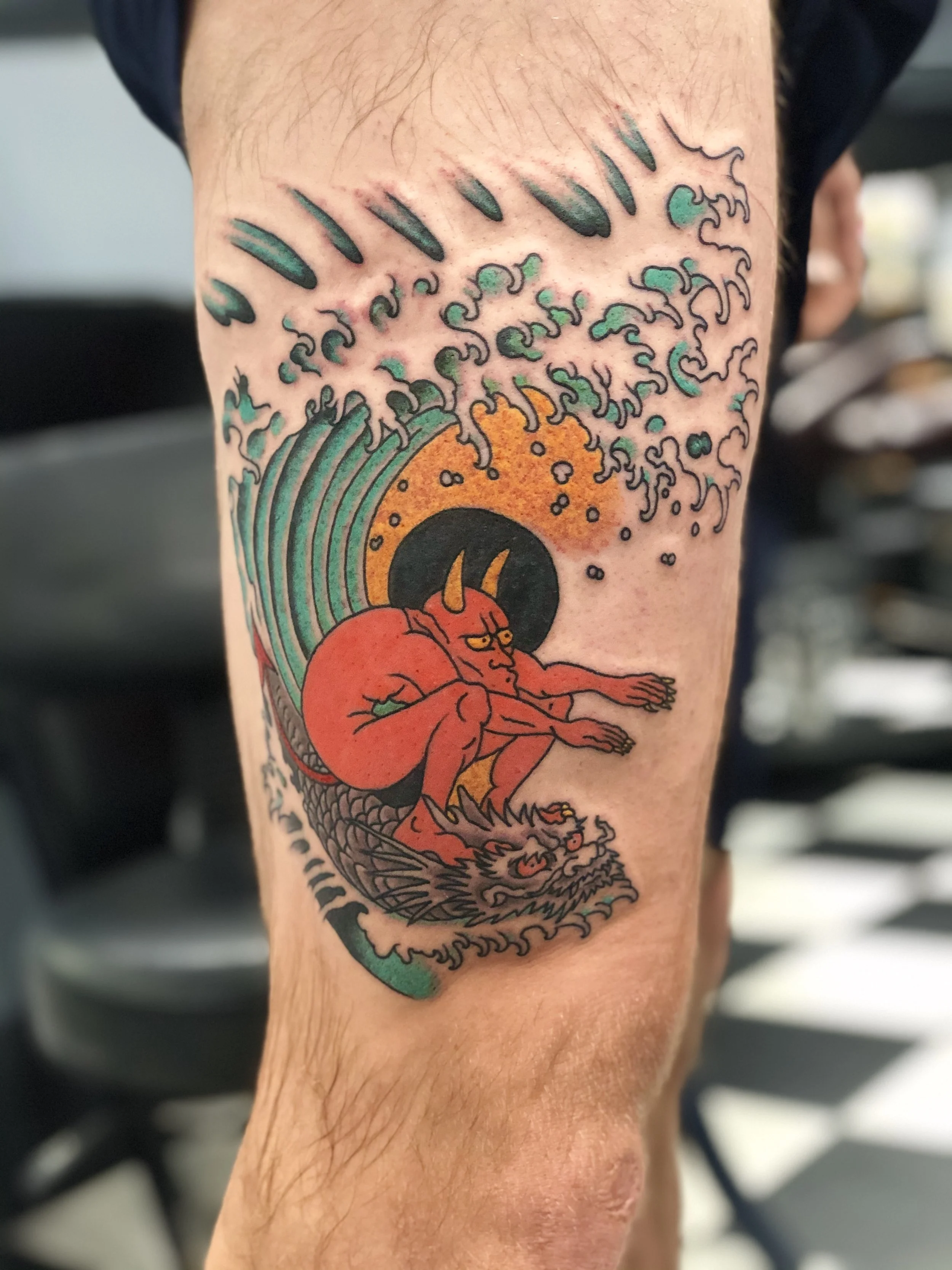 Japanese-influenced color leg tattoo featuring a red horned devil surfing a cresting teal wave atop a dragon, with a bold golden yellow sun and dynamic water spray, inspired by Ed Hardy's classic Surfin' Satan painting in a  Japanese traditional fusi