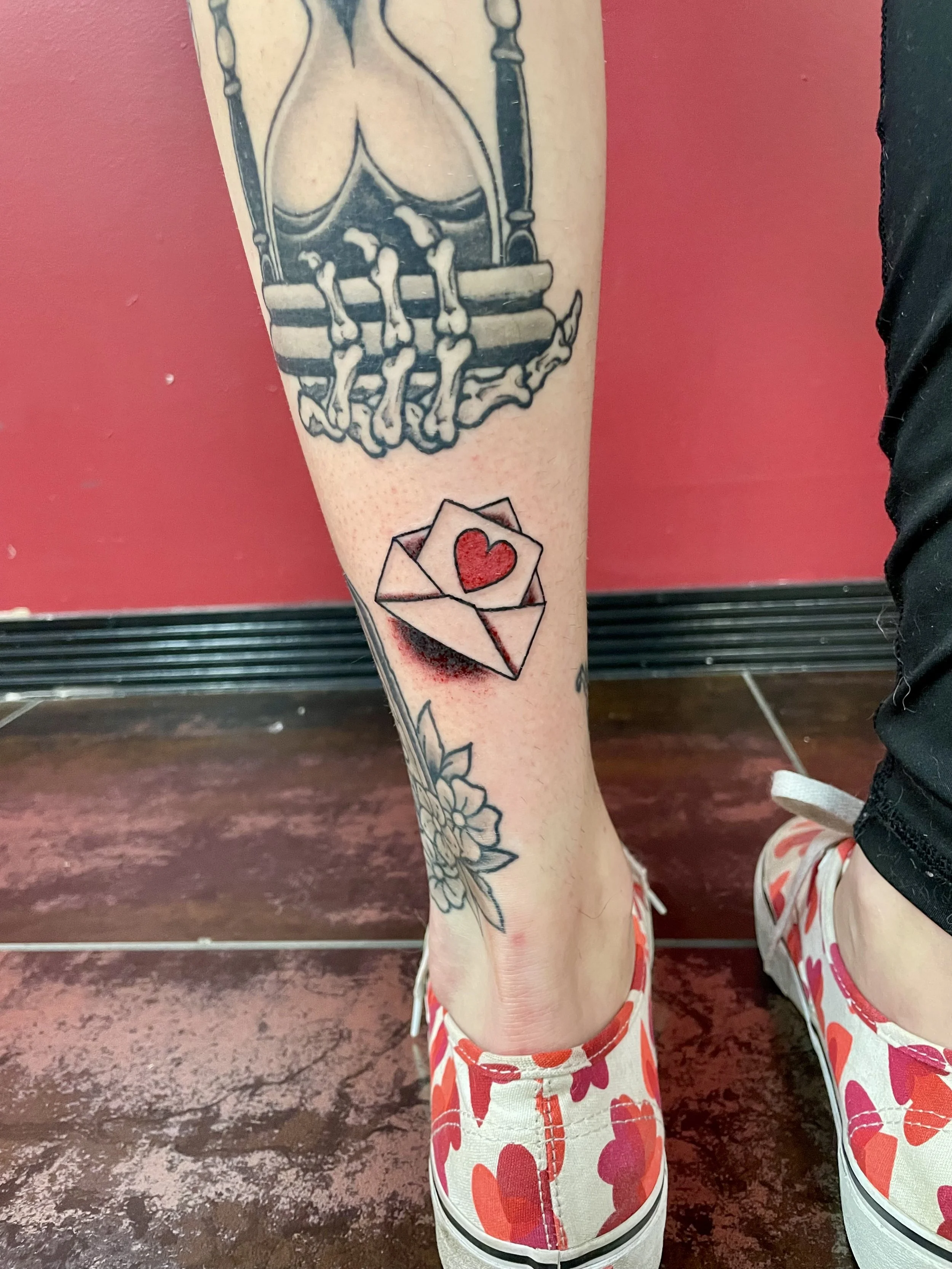 Traditional American color ankle tattoo featuring a small open envelope with a red heart inside, set among existing leg work including a skeleton hourglass above and floral designs below, custom tattoo by Nick at Big Kahuna Tattoo in Boca Raton, FL.