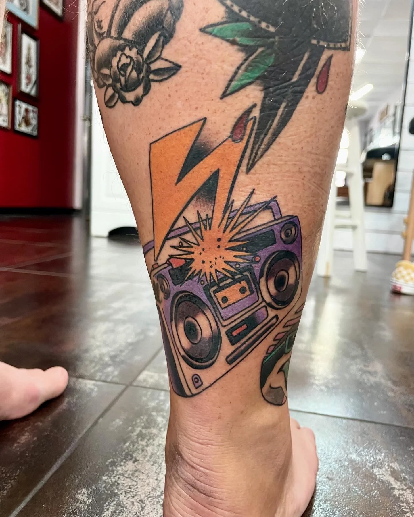 Color leg sleeve tattoo featuring a bold purple boombox with a cassette tape and exploding speaker graphic, struck by a large orange Bad Brains-inspired lightning bolt, set among existing sleeve work, custom tattoo by Nick at Big Kahuna Tattoo in Boc