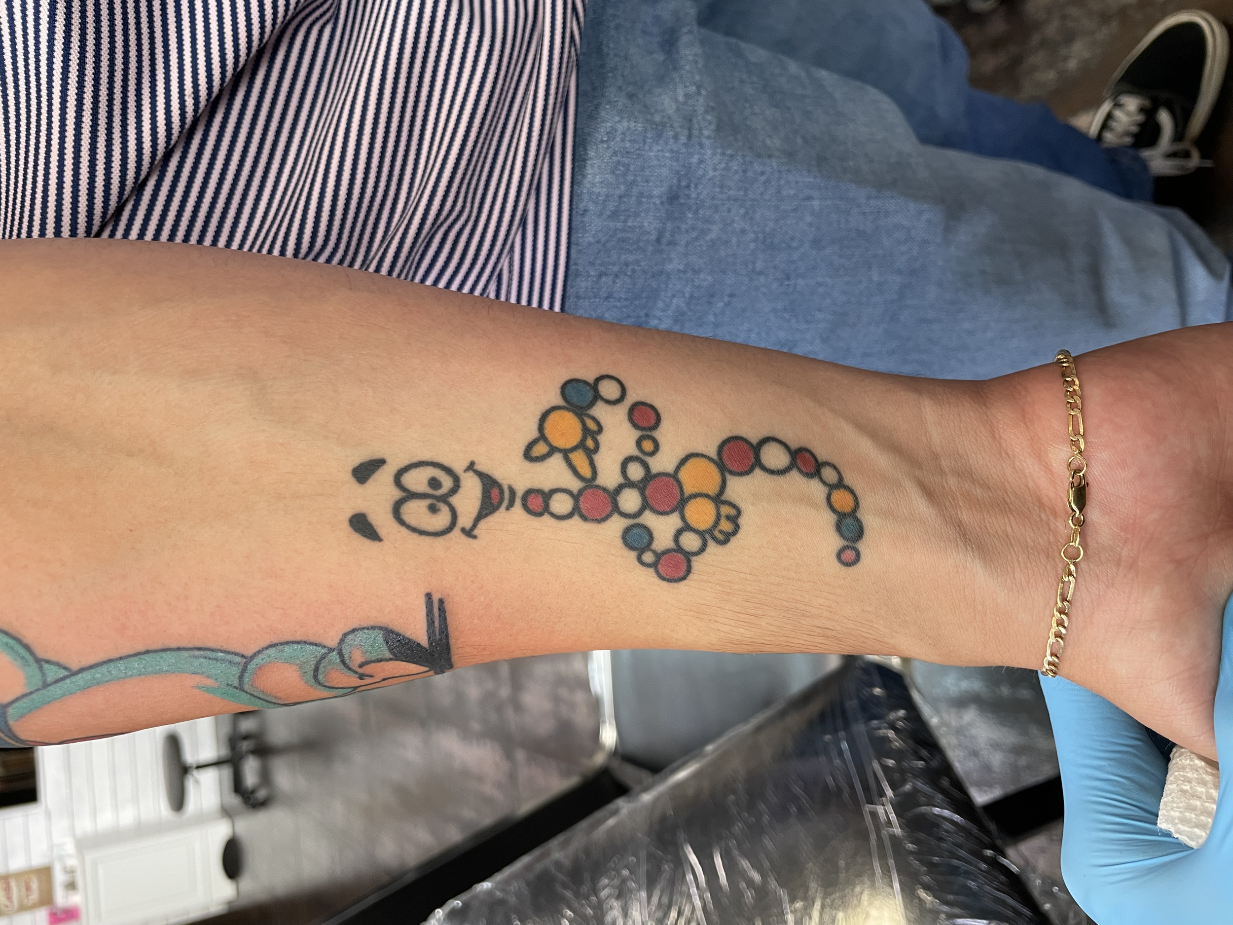 Color forearm tattoo featuring Mr. DNA, the beloved cartoon character from the 1993 Jurassic Park movie, depicted as a smiling double helix figure made of multicolored circles, custom pop culture tribute tattoo by Nick at Big Kahuna Tattoo in Boca Ra